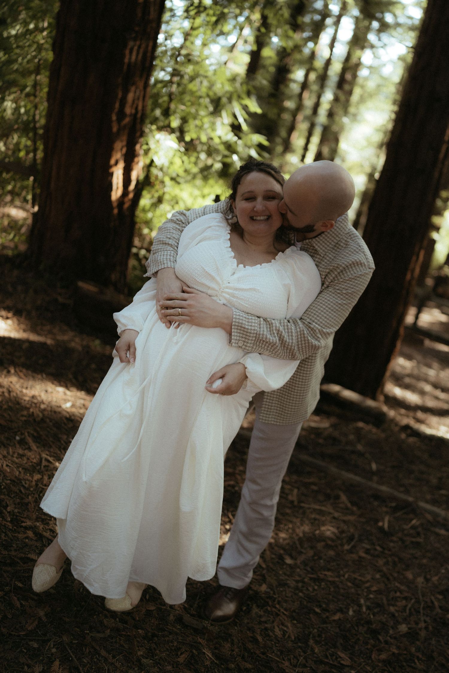 A romantic forest photoshoot featuring a couple in light-colored clothing among towering trees.