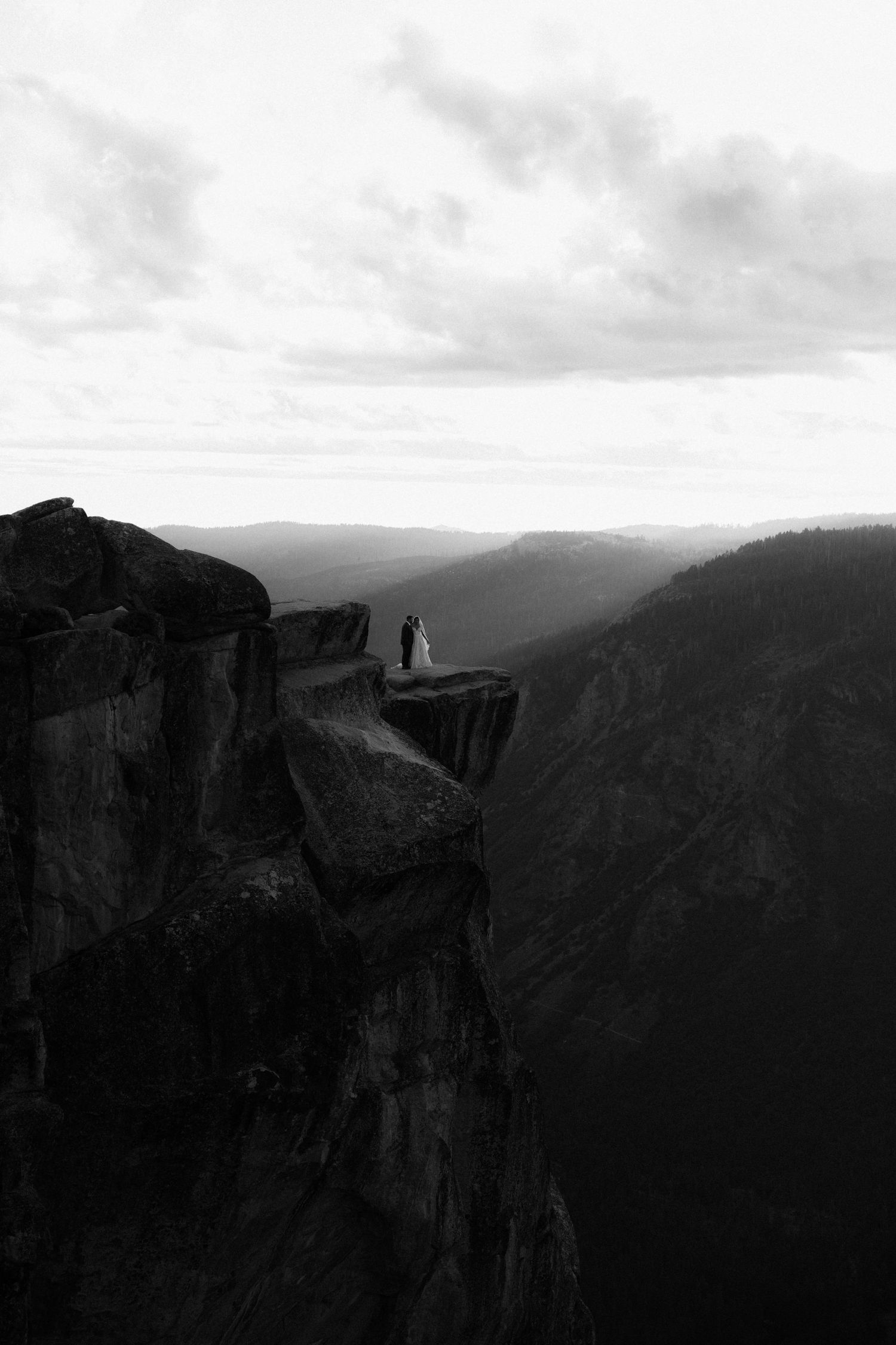 Bride and groom stand on dramatic cliff edge at Yosemite's Taft Point in stunning black and white wedding photo.