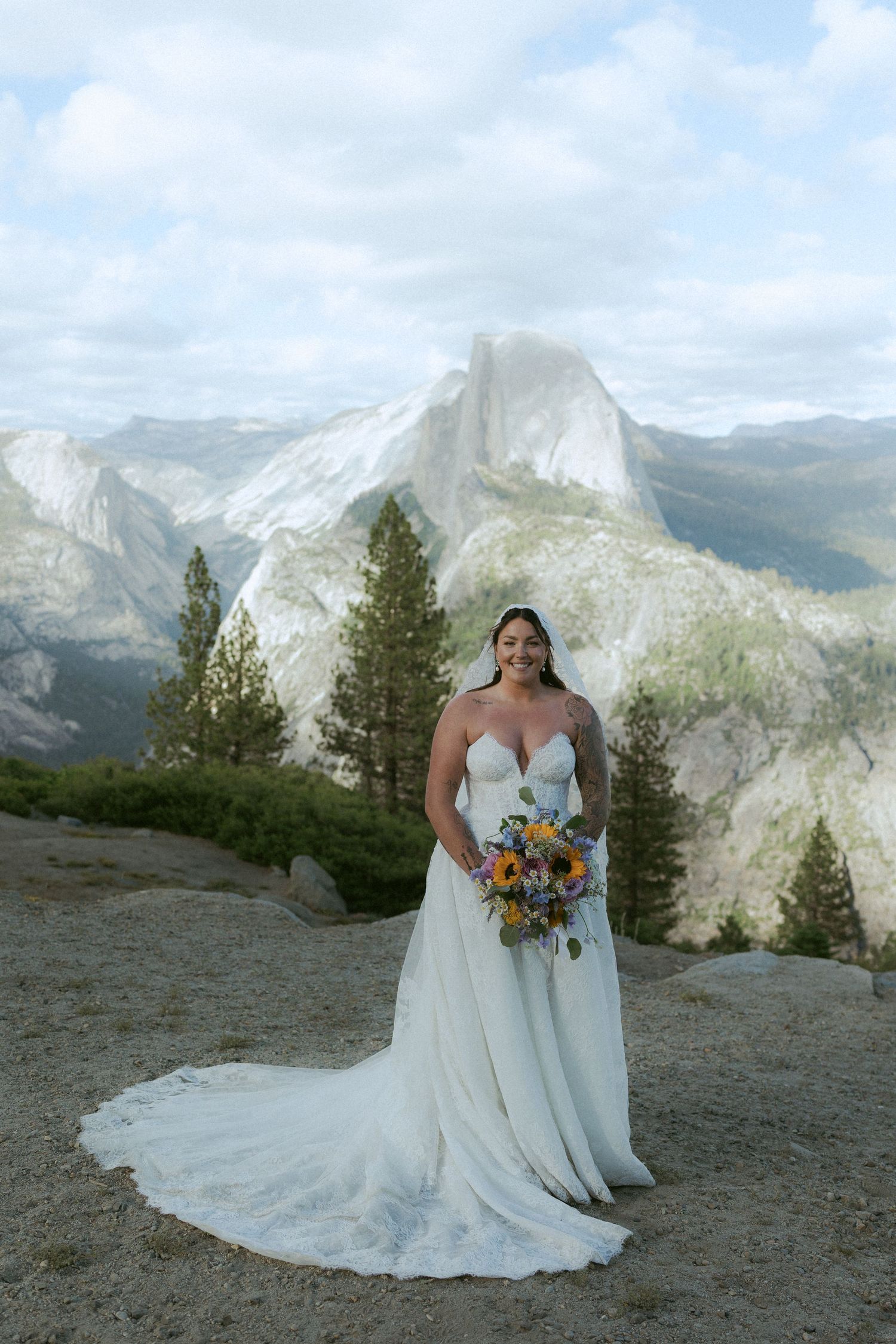 Smiling bride in strapless gown holds colorful bouquet with Half Dome Yosemite view.
