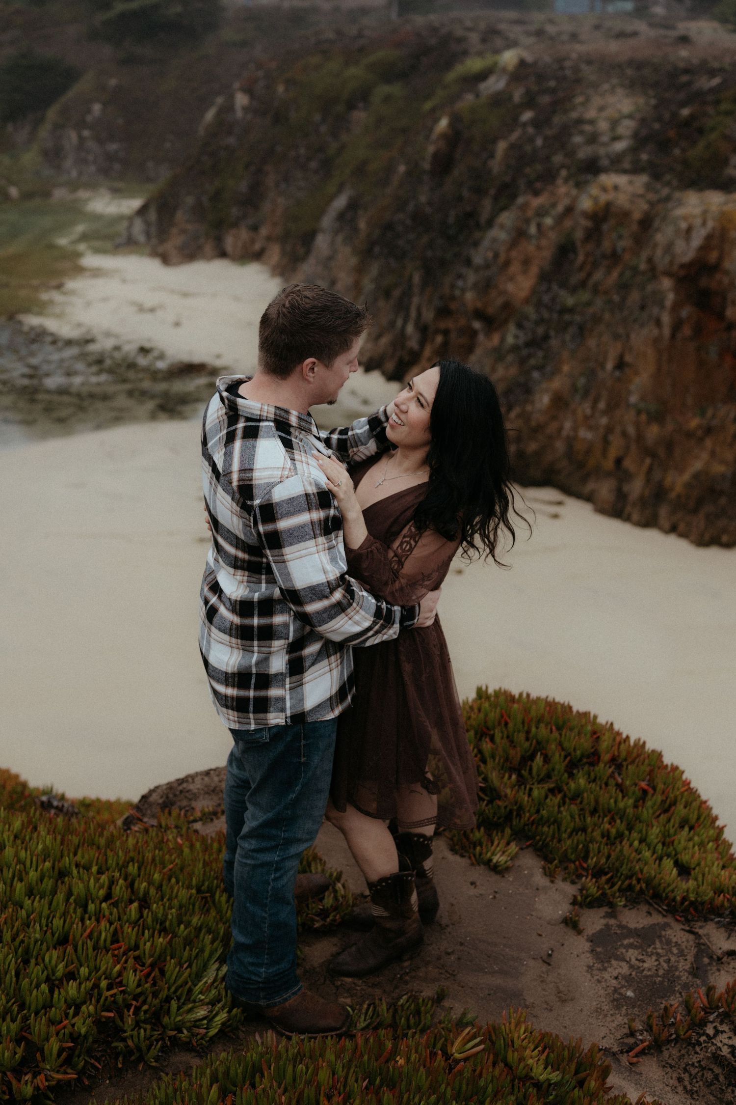 Couple embracing on rugged coastal cliffs with native vegetation in the foreground.