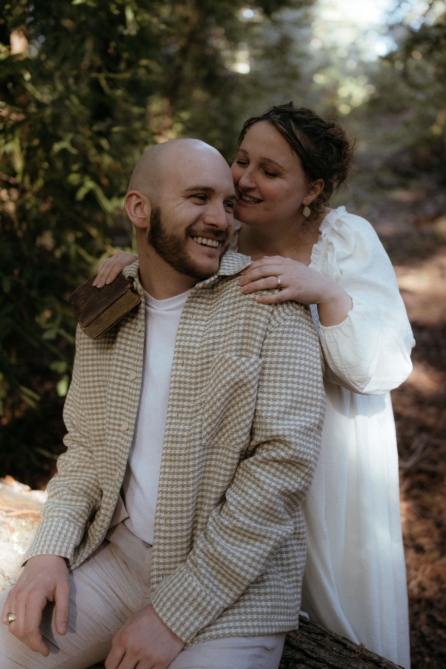 A couple shares intimate moments in a forest setting wearing light colored clothing.