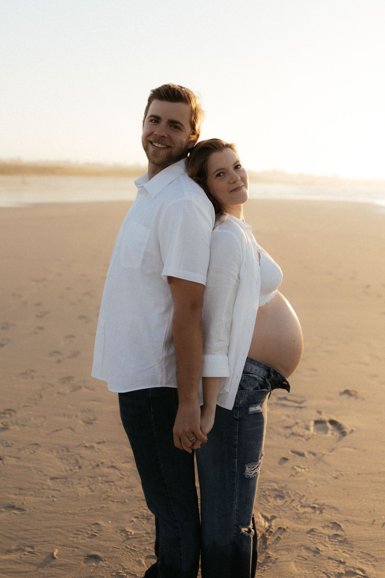 Couple in white shirts standing together on sandy beach during golden hour.