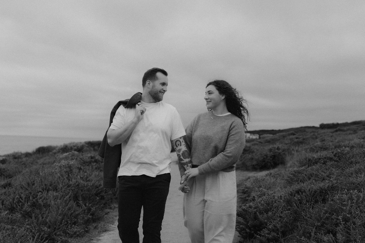 Two people walking together along a scenic coastal path with hills and ocean views in the background.