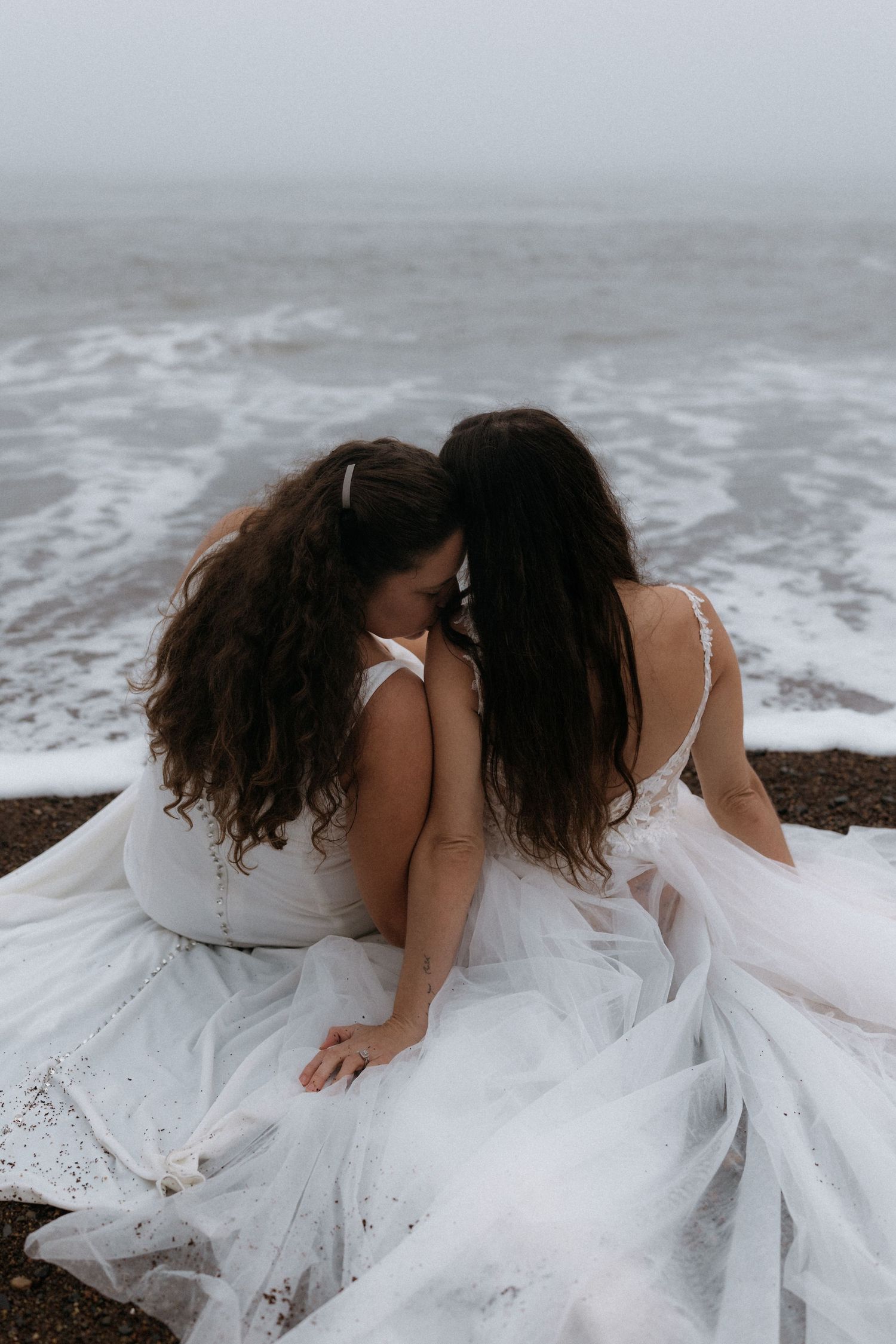 Two people in white wedding dresses lay on a beach with waves crashing behind them in a romantic pose.