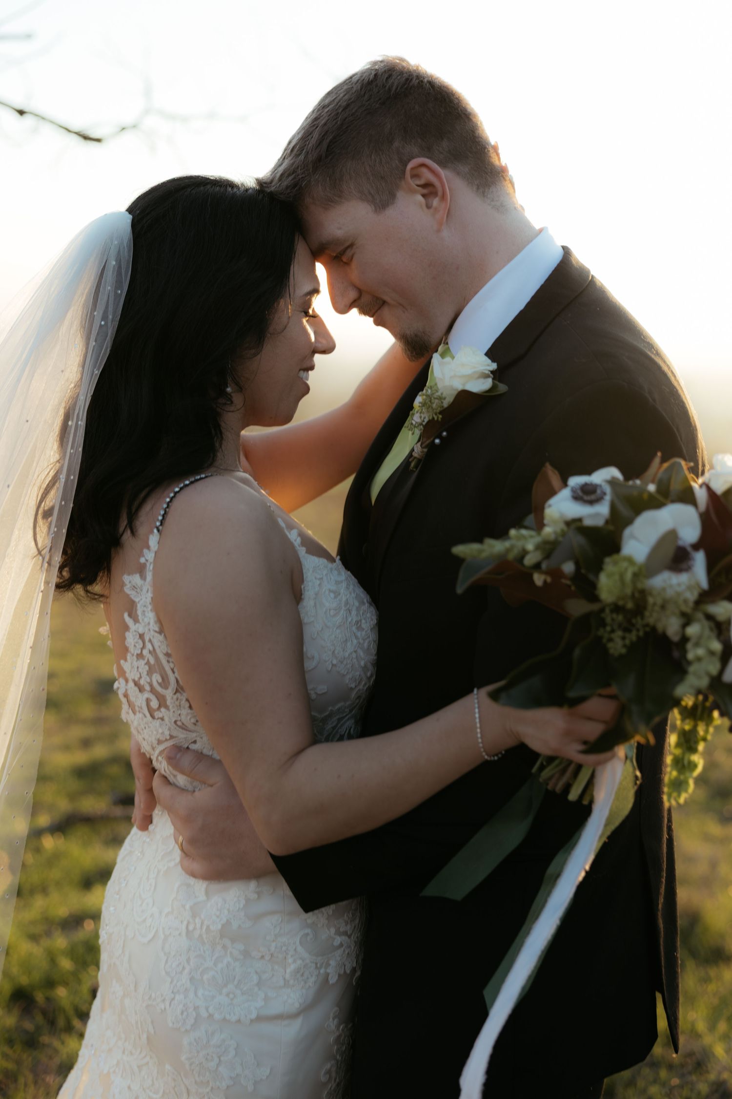 Tender moment between newlyweds as they embrace during sunset with bouquet in hand.