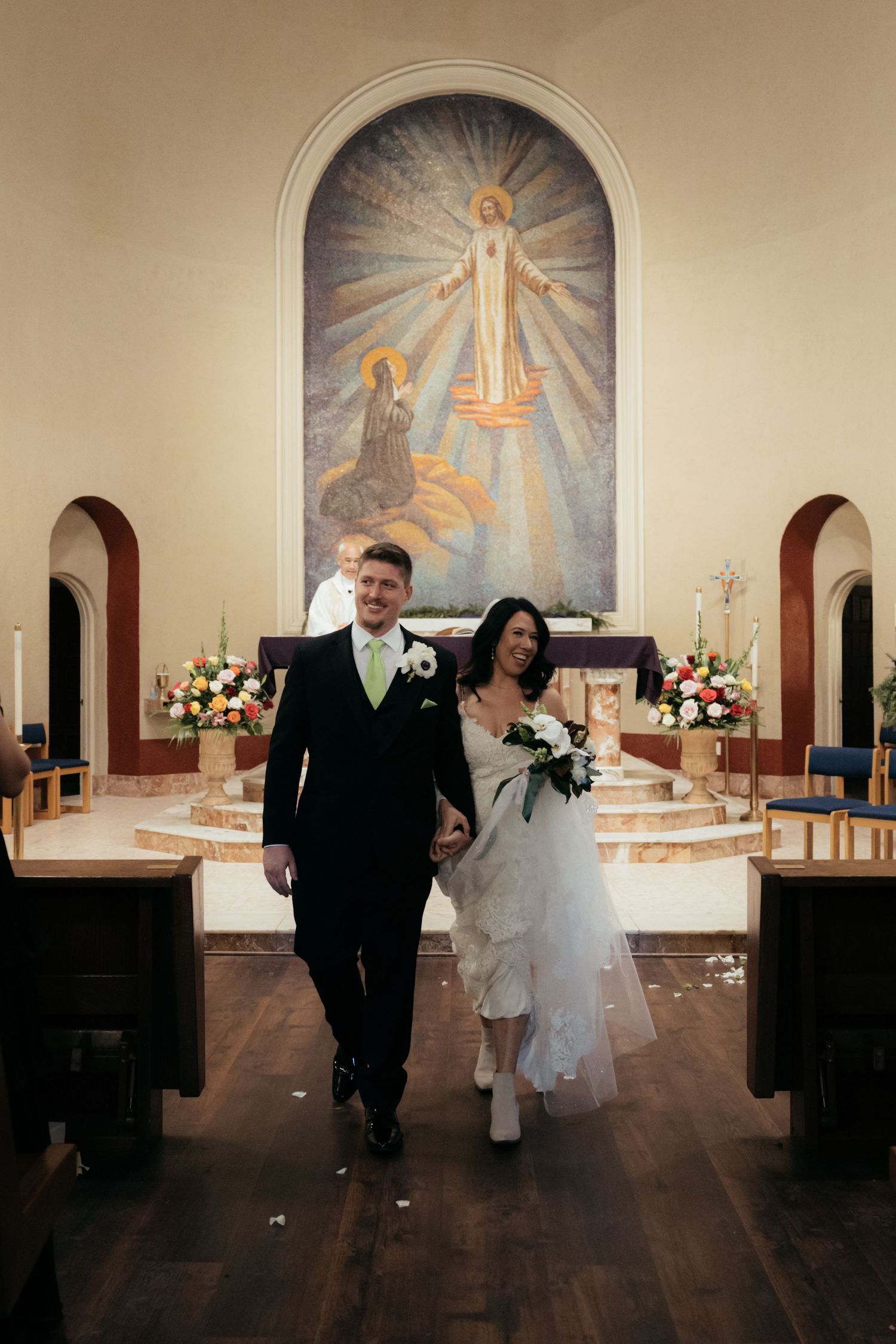 A couple walks down the church aisle with a religious mural visible in the background.