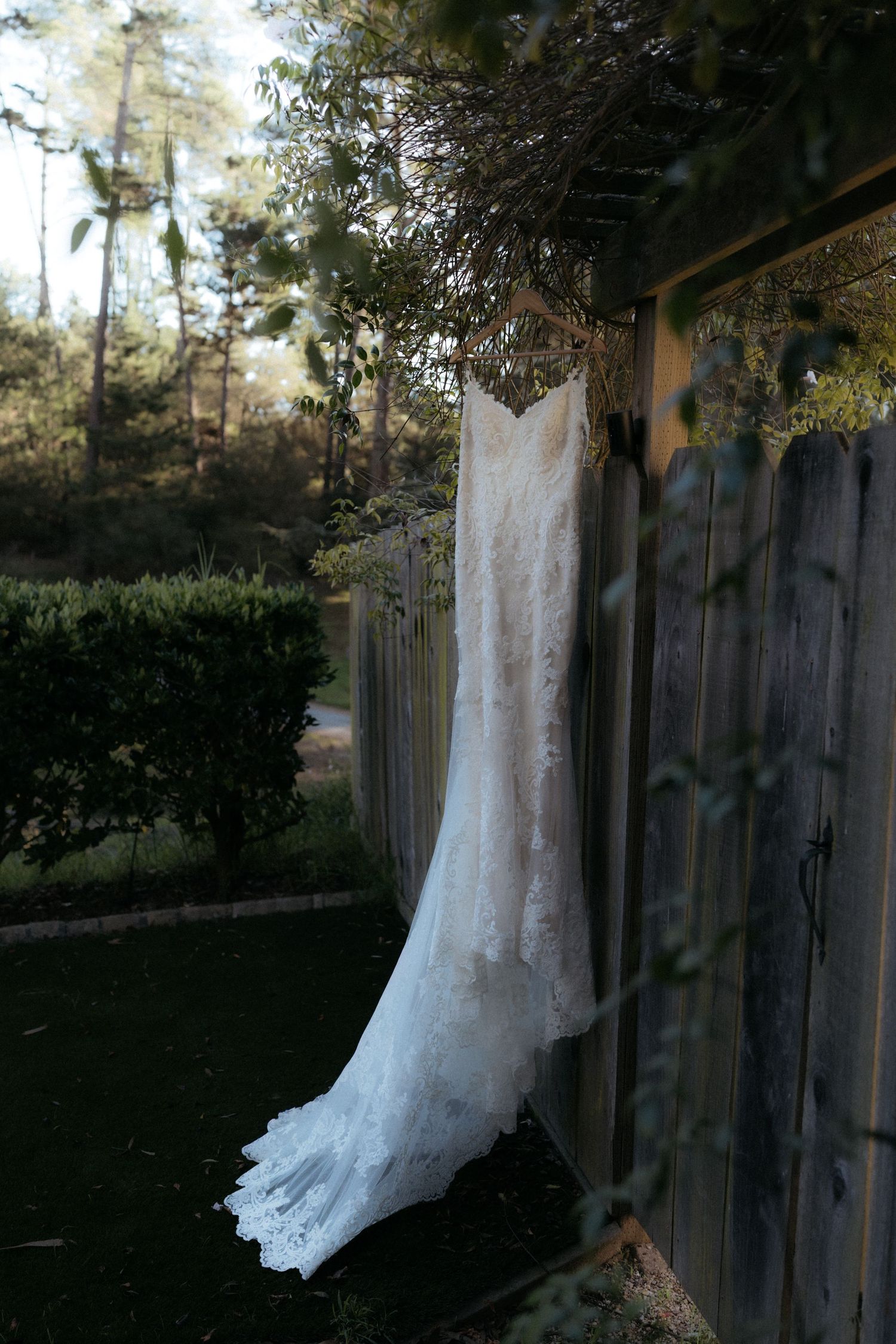 A white lace wedding dress hangs elegantly on a wooden fence outdoors with trees and greenery in the background.