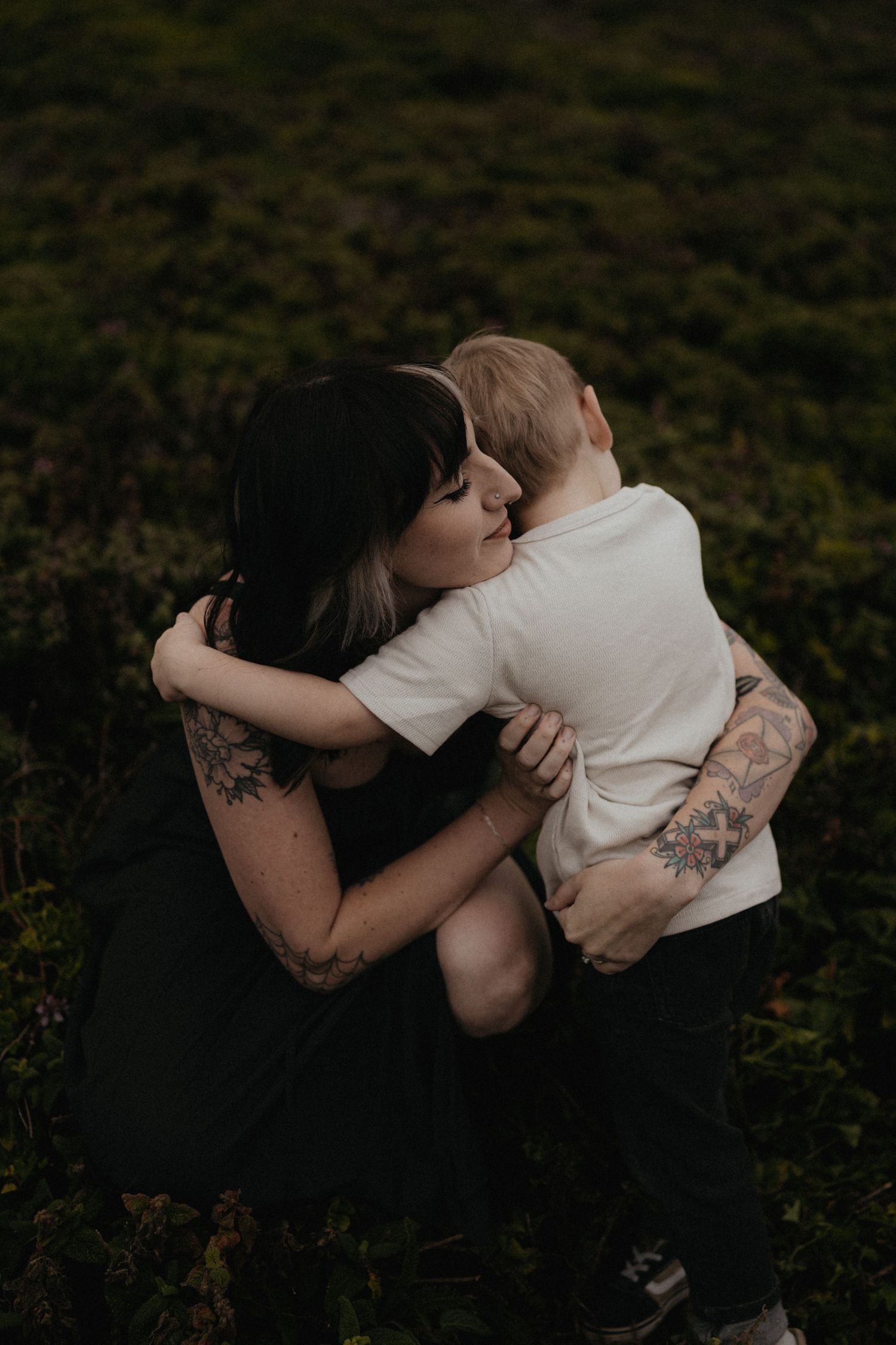 A tattooed mother hugs her young child in a lush green outdoor setting, sharing a tender moment.