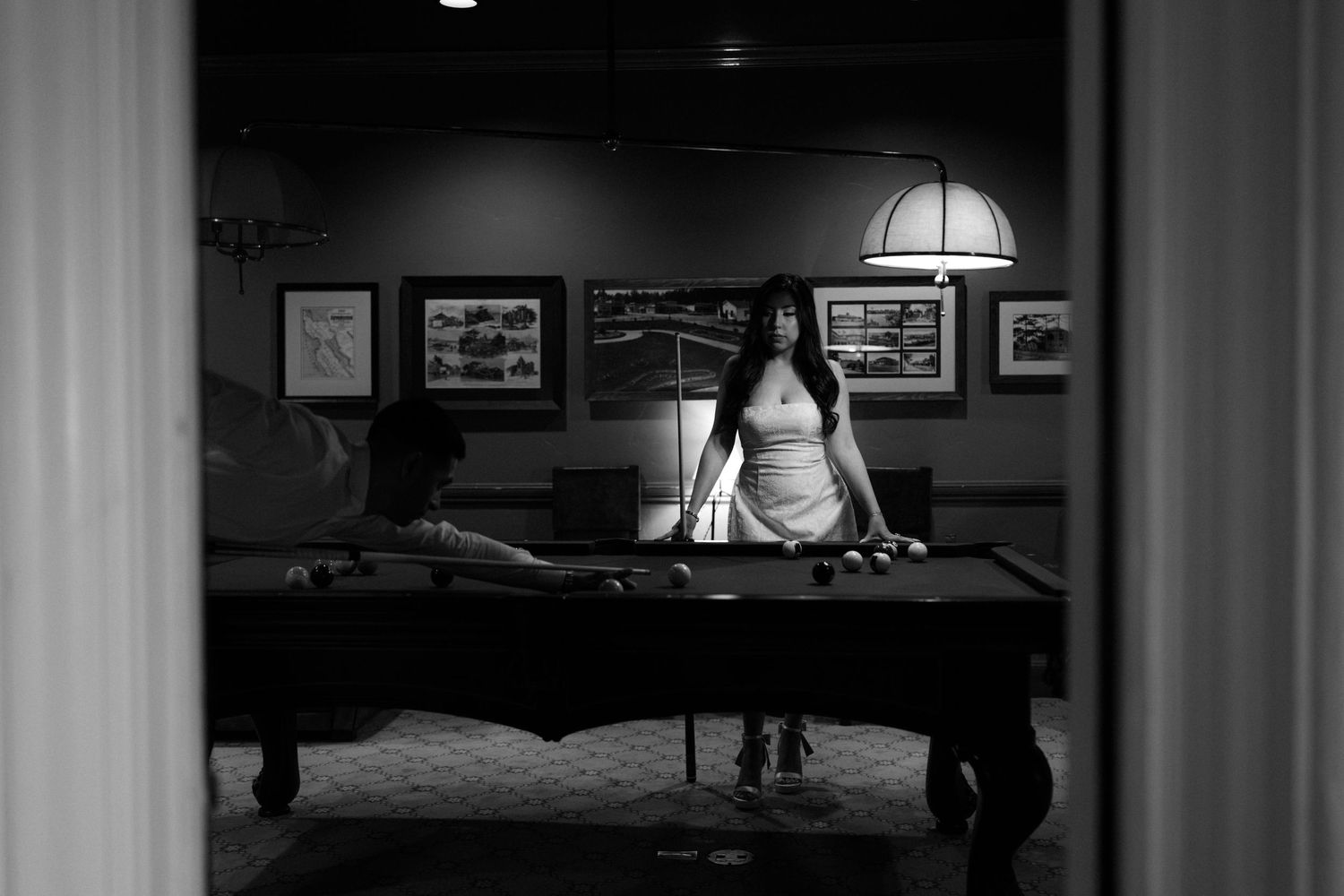 Black and white photo of a woman standing by a pool table in a dimly lit billiards room with framed art on walls.