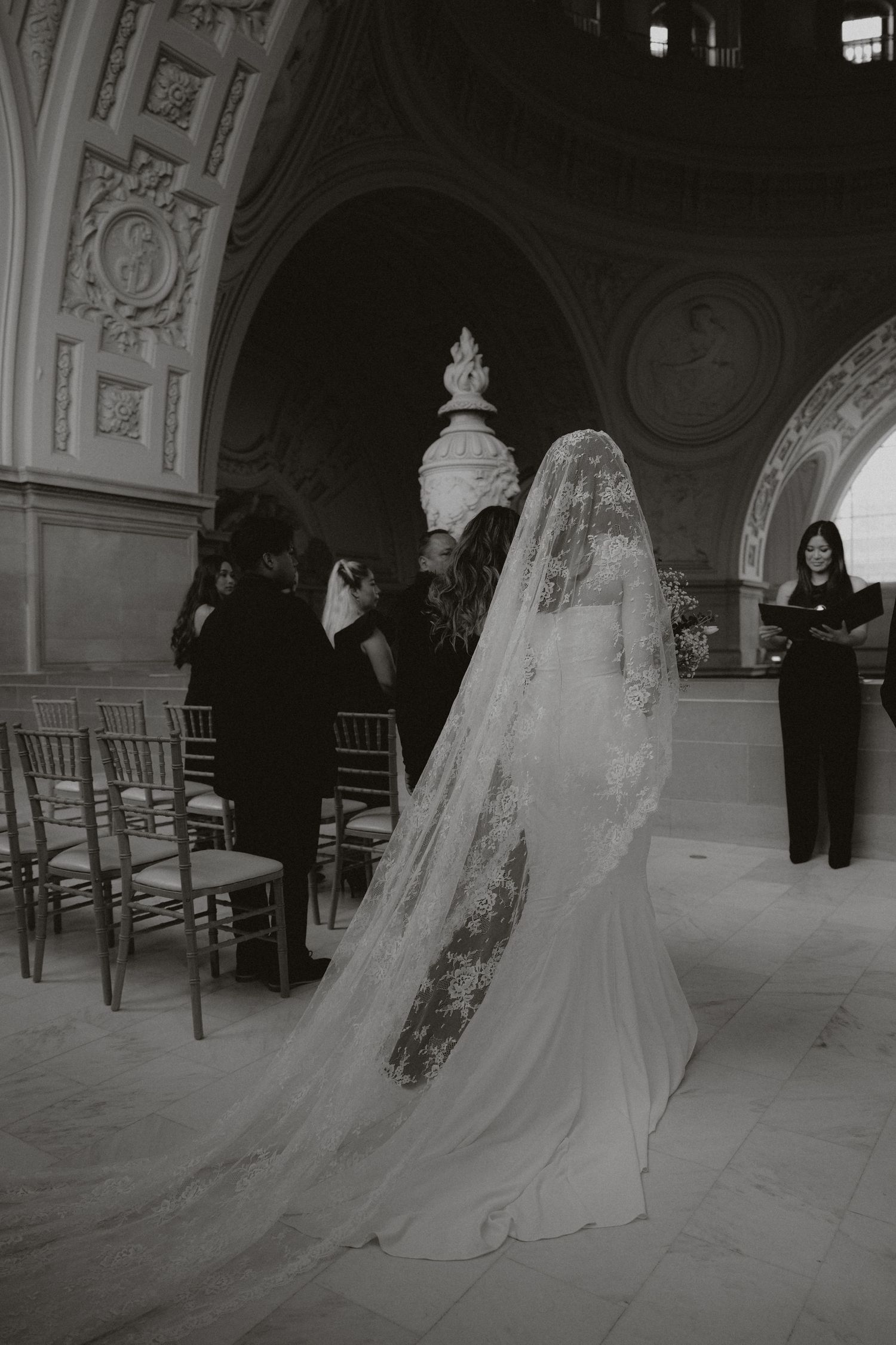 A bride in a flowing veil walks through an ornate hallway with classical architectural elements.