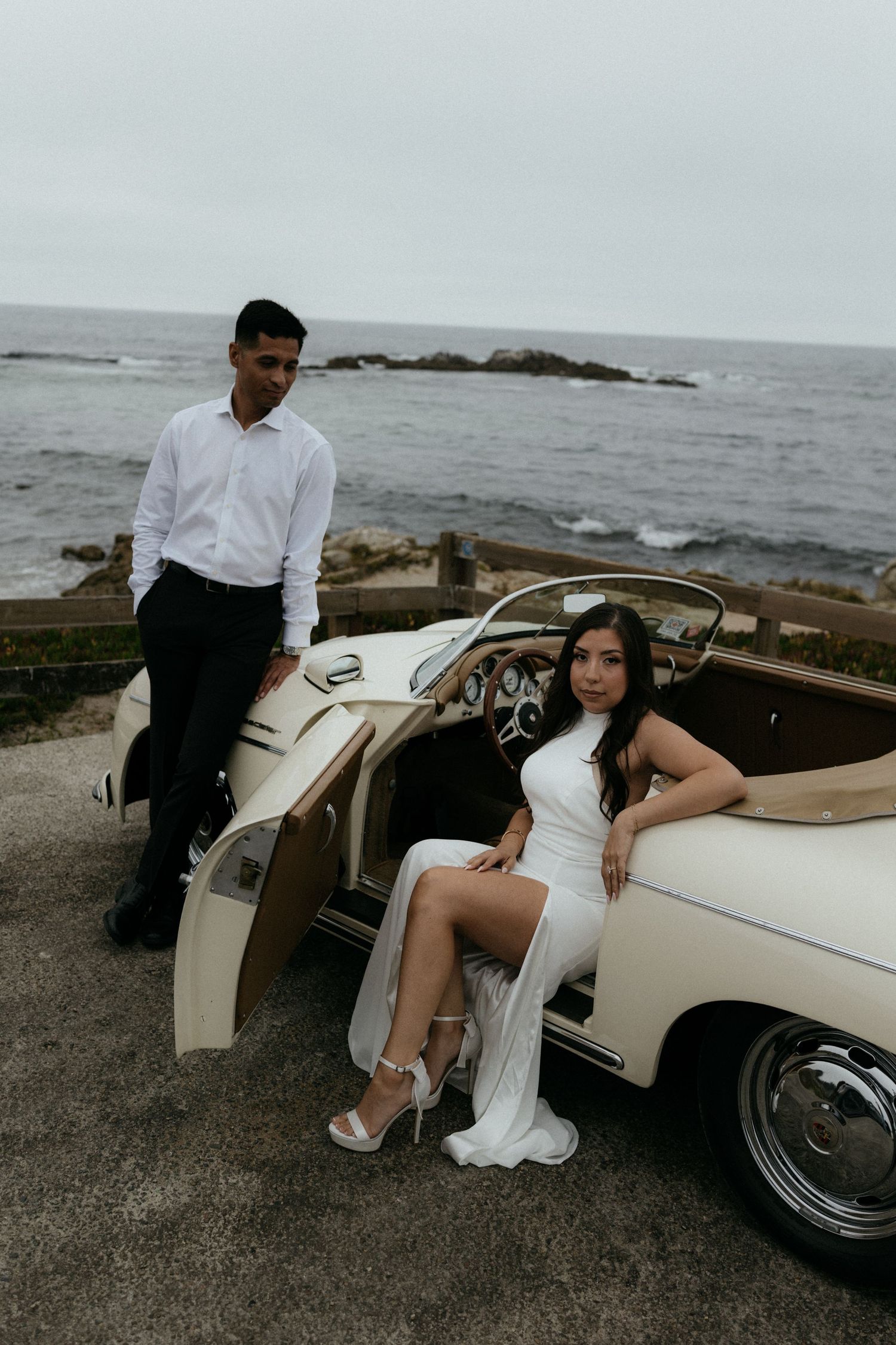 Bride in white gown posing by vintage cream convertible with groom by ocean coastline on overcast day.