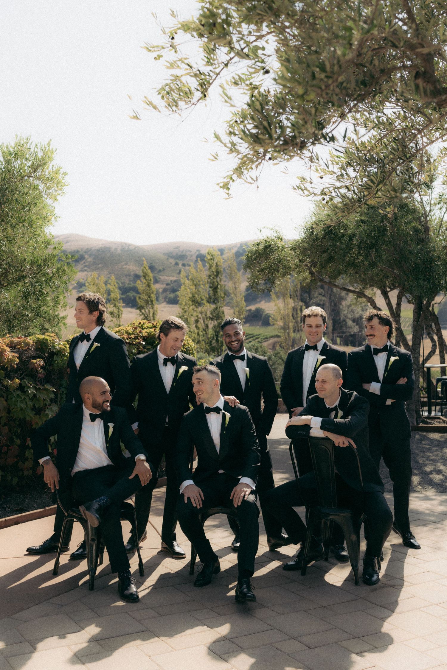 Groomsmen in black tuxedos sit casually on stone wall at outdoor wedding venue with olive trees in background.