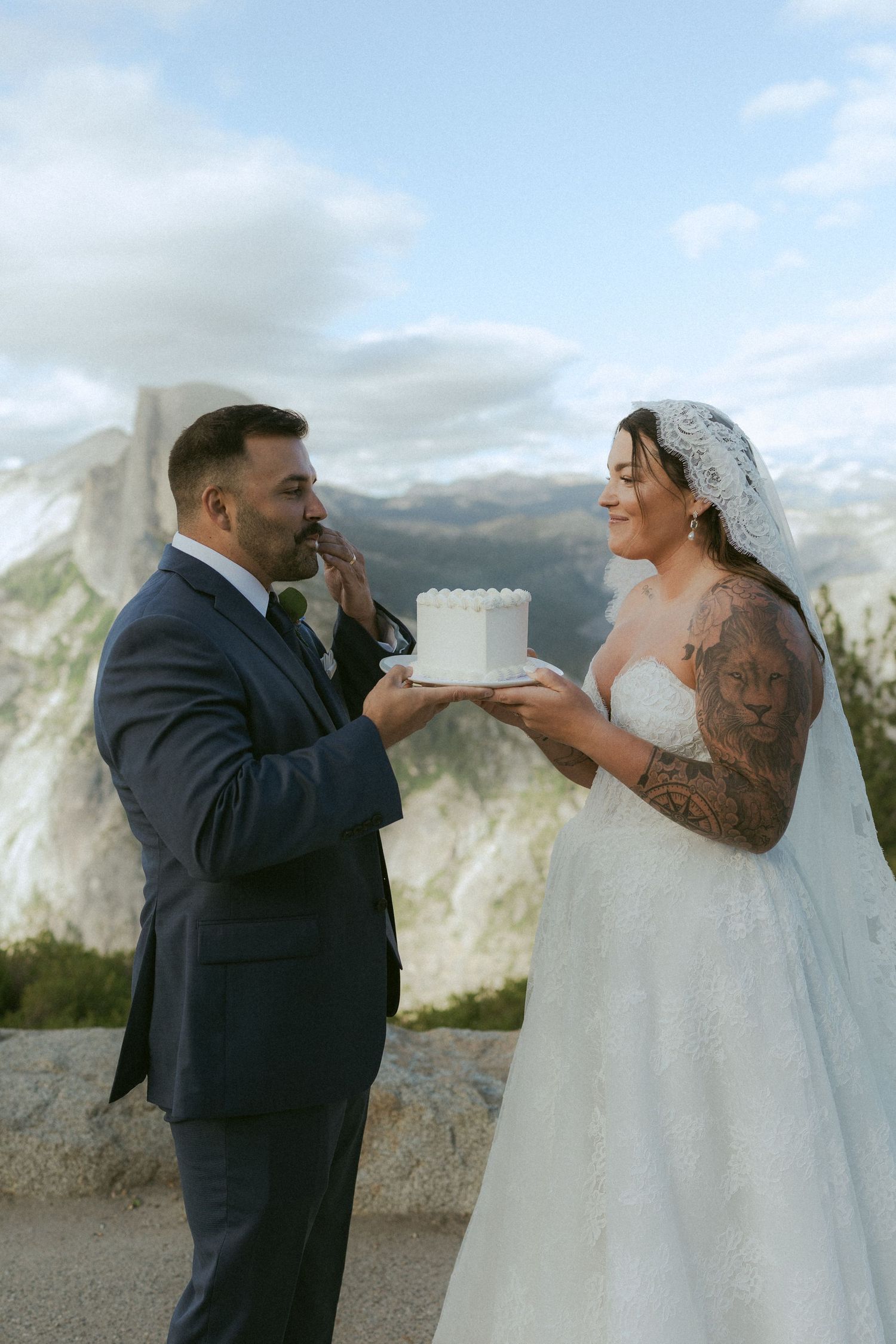 Bride feeds groom wedding cake at Yosemite overlook with Half Dome in background during outdoor elopement.