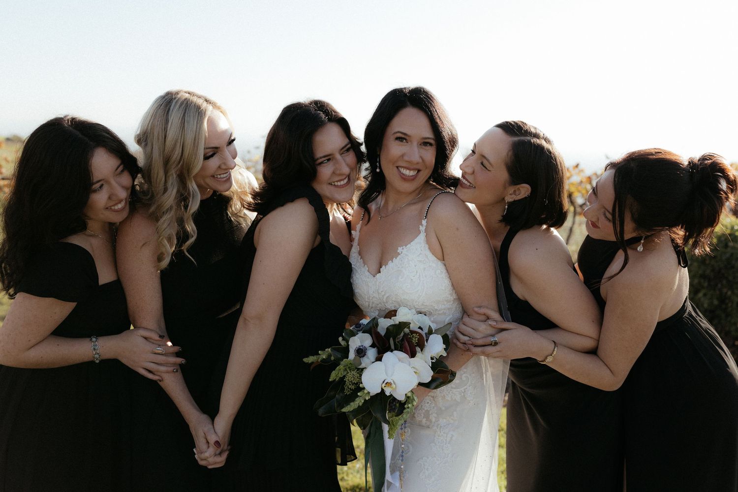 Bridesmaids in black dresses gather around bride in white gown holding bouquet during outdoor wedding celebration.