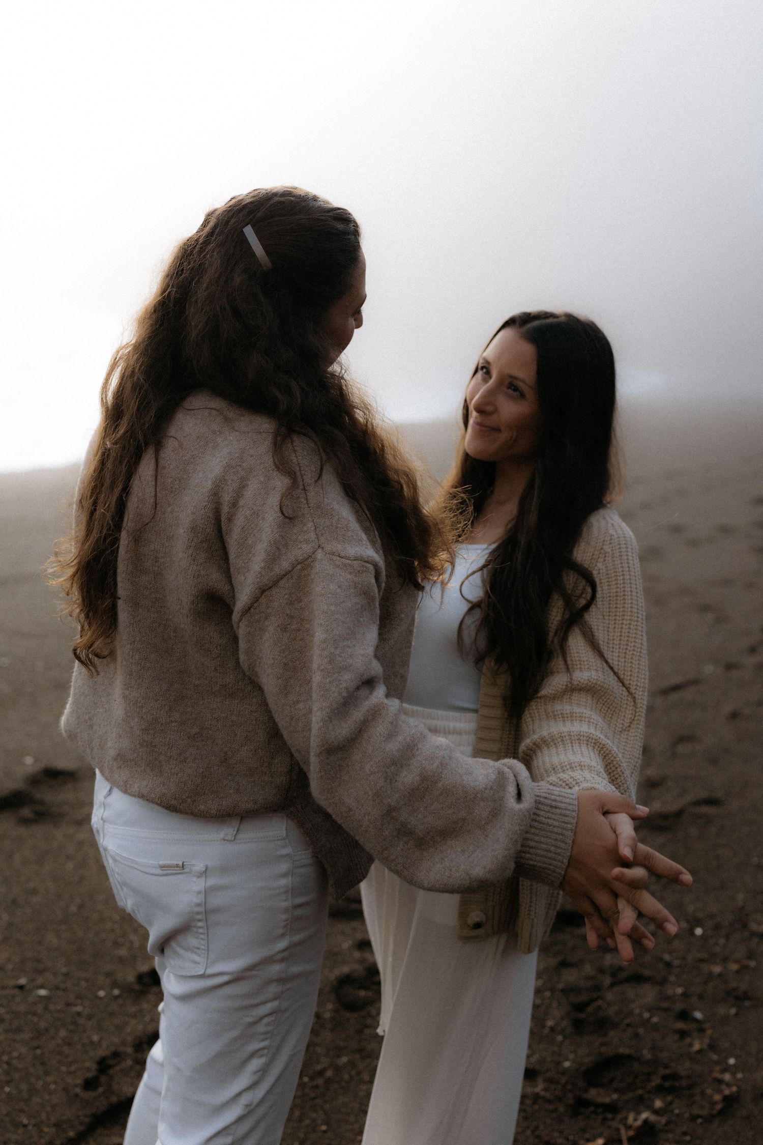 Two people in cozy sweaters share an intimate moment on a misty beach in neutral earth tones.