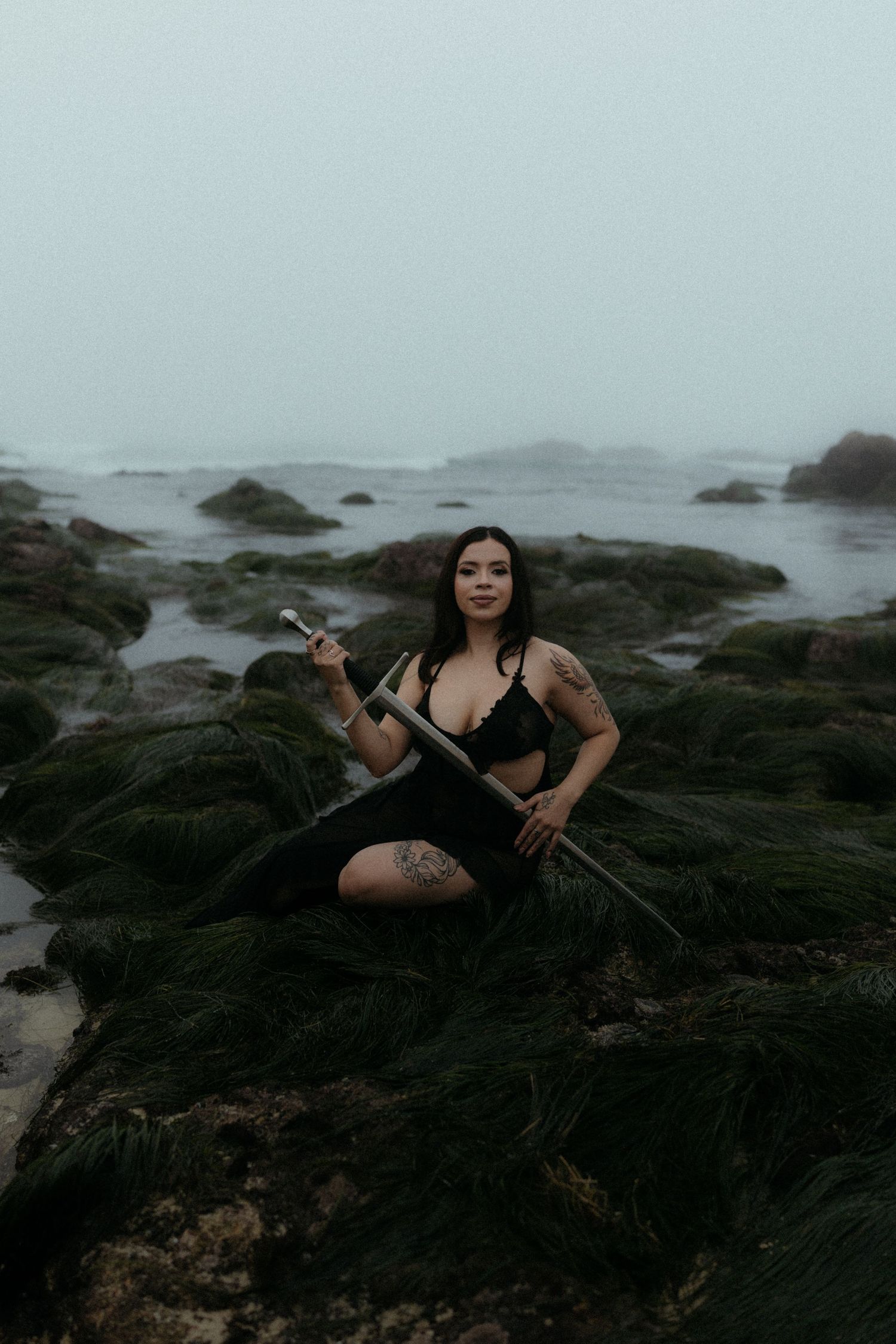 Dramatic photoshoot on rocky coastal terrain with dark moody atmosphere and misty ocean backdrop.