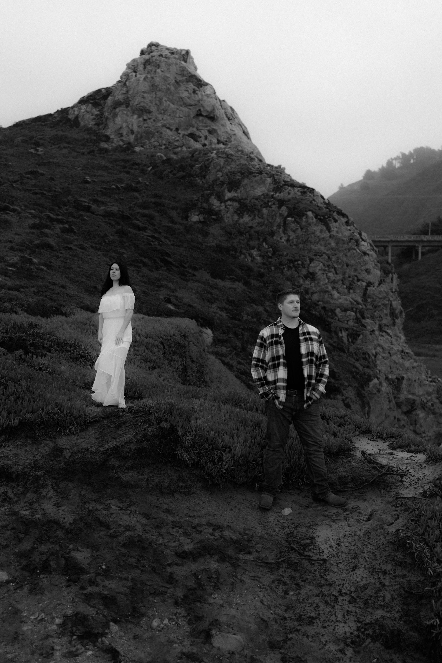 Couple in white dress and plaid shirt posing against dramatic rocky hillside.