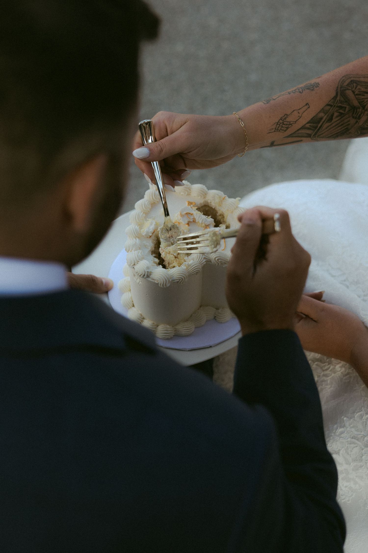 Tattooed bride and groom cut small white wedding cake together during outdoor elopement celebration.