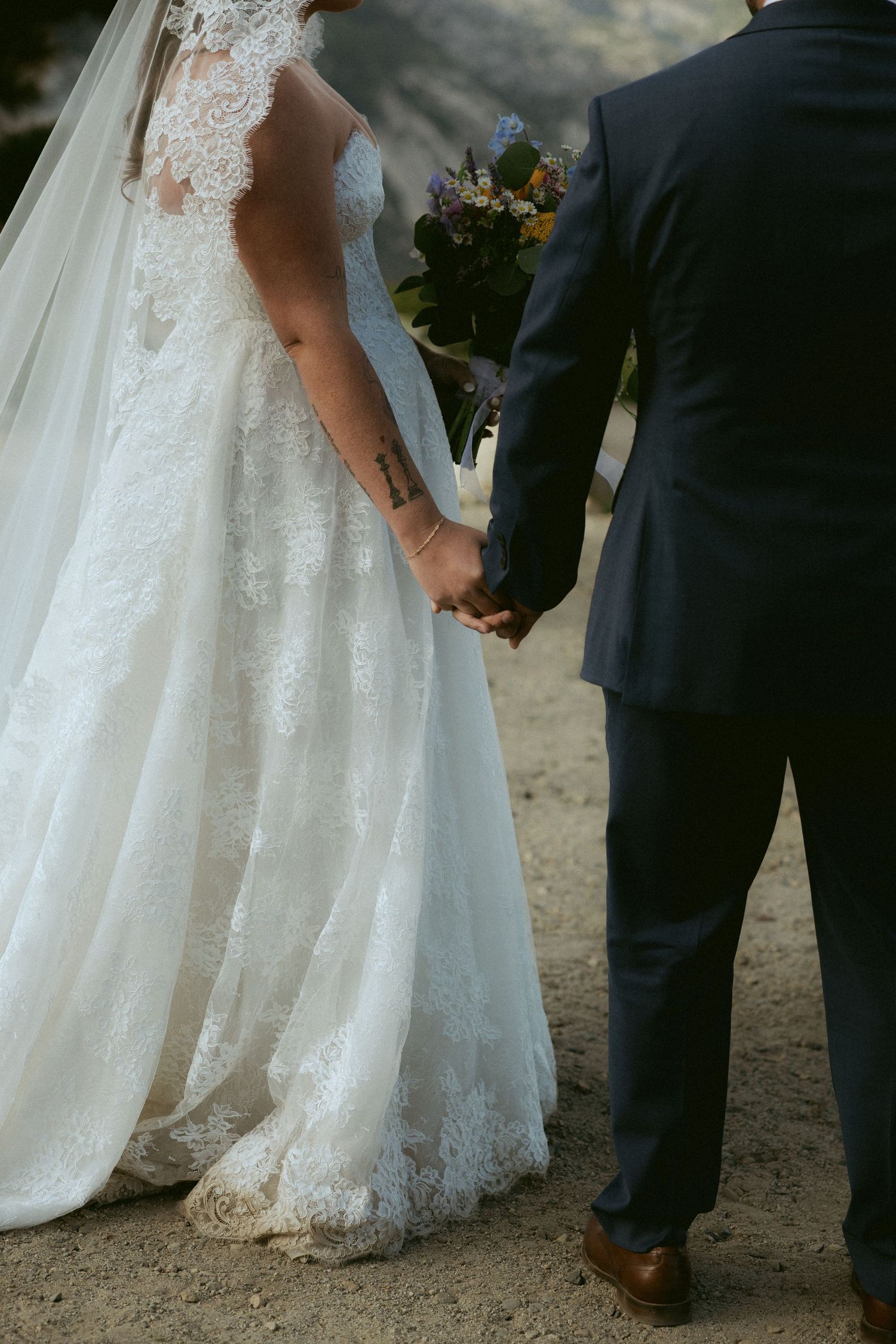 Bride in lace wedding gown holding colorful bouquet while holding grooms hand outdoors.