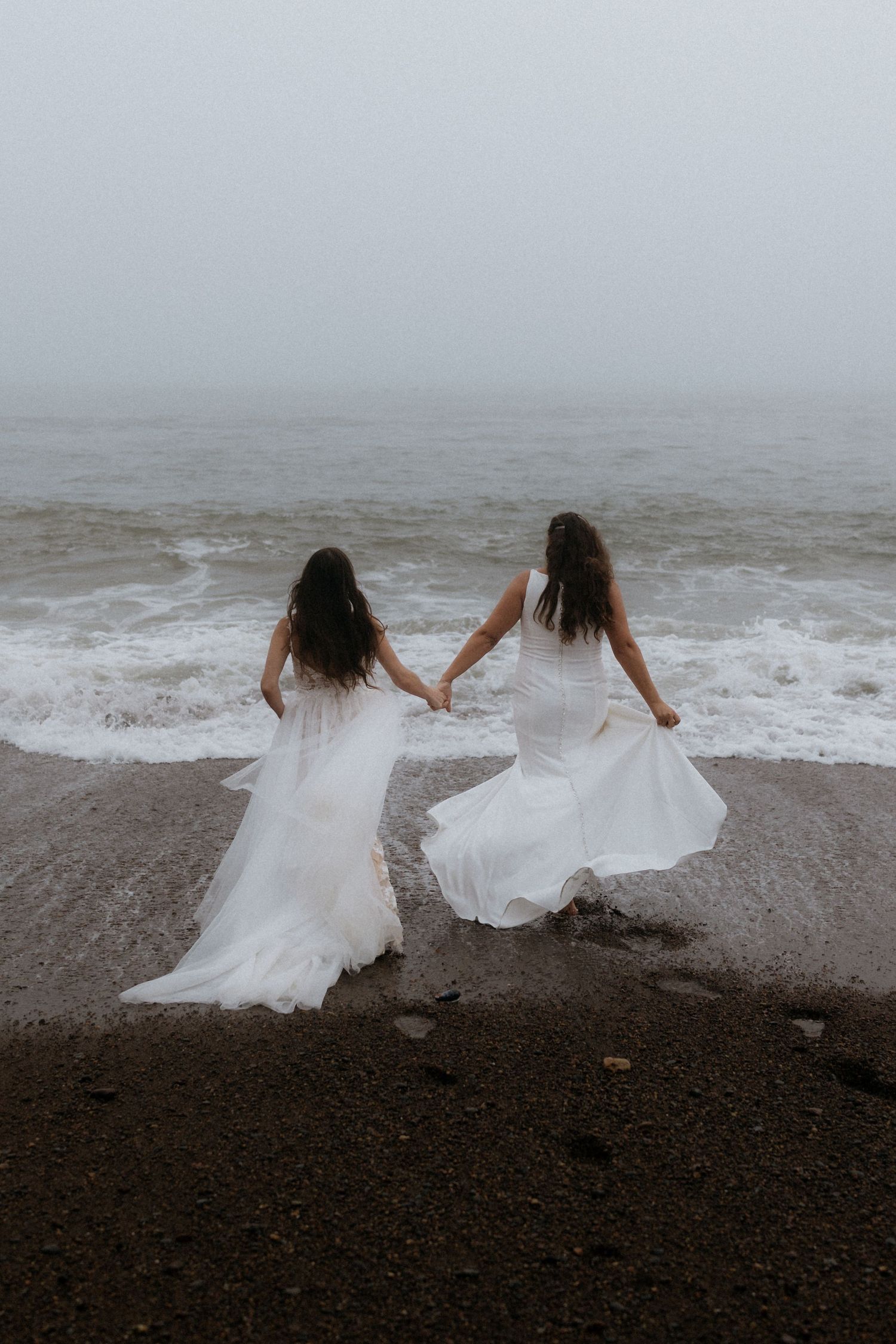 Two figures in flowing white dresses hold hands while standing at the edge of a misty beach with crashing waves.