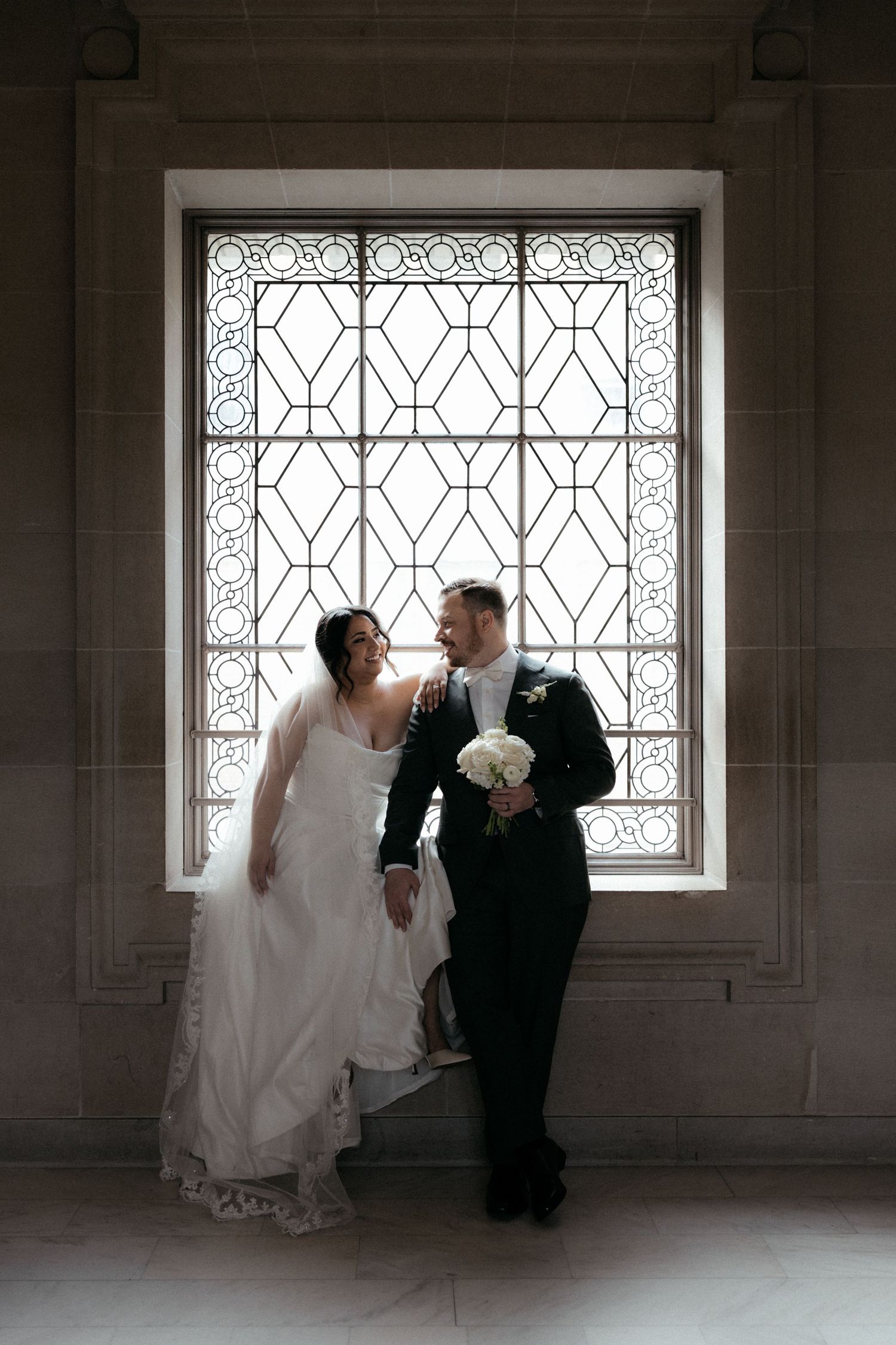 A couple in wedding attire pose together in front of an ornate window with geometric iron detailing.