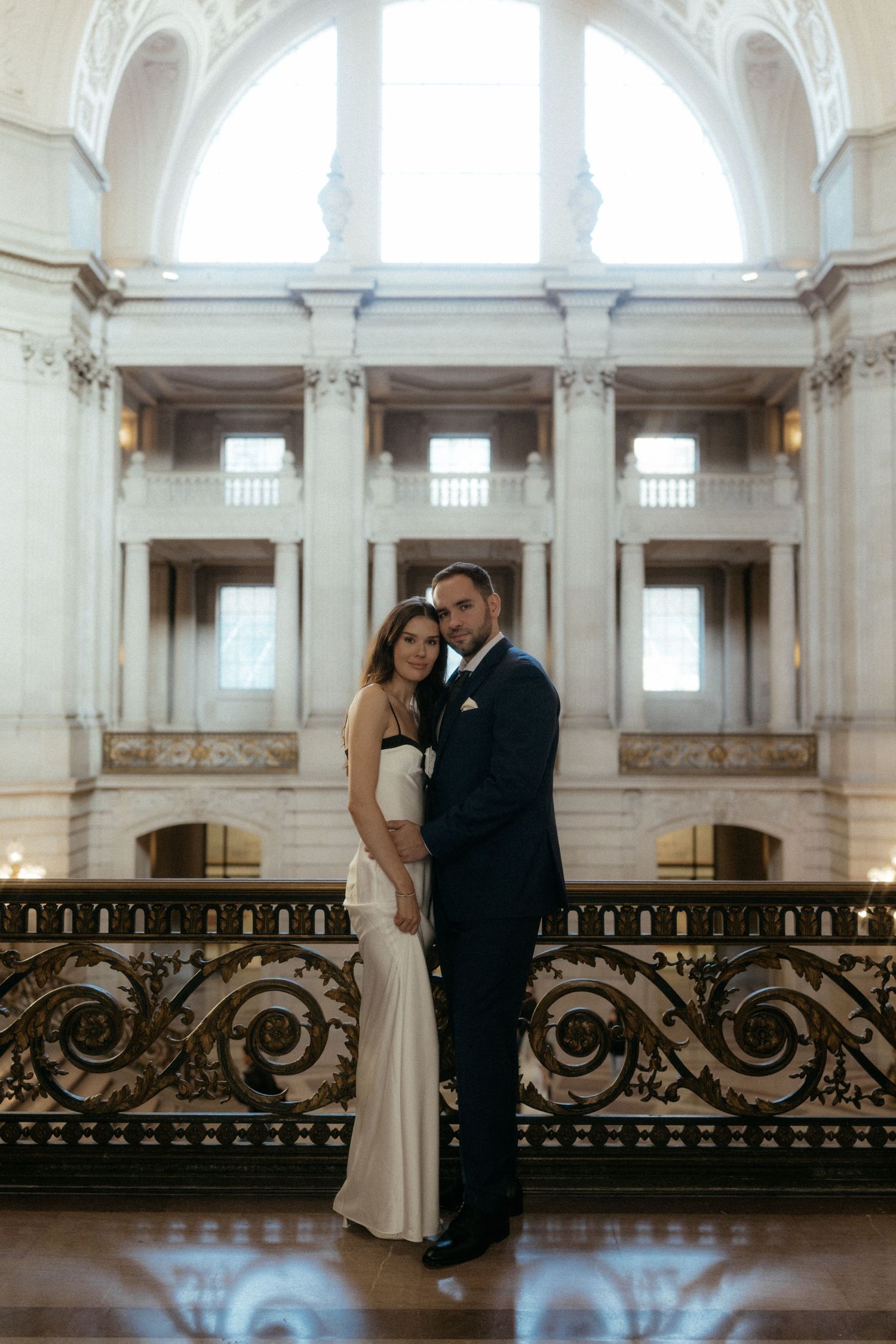 A romantic couple shares an intimate moment in a grand historic building with ornate railings and natural light.