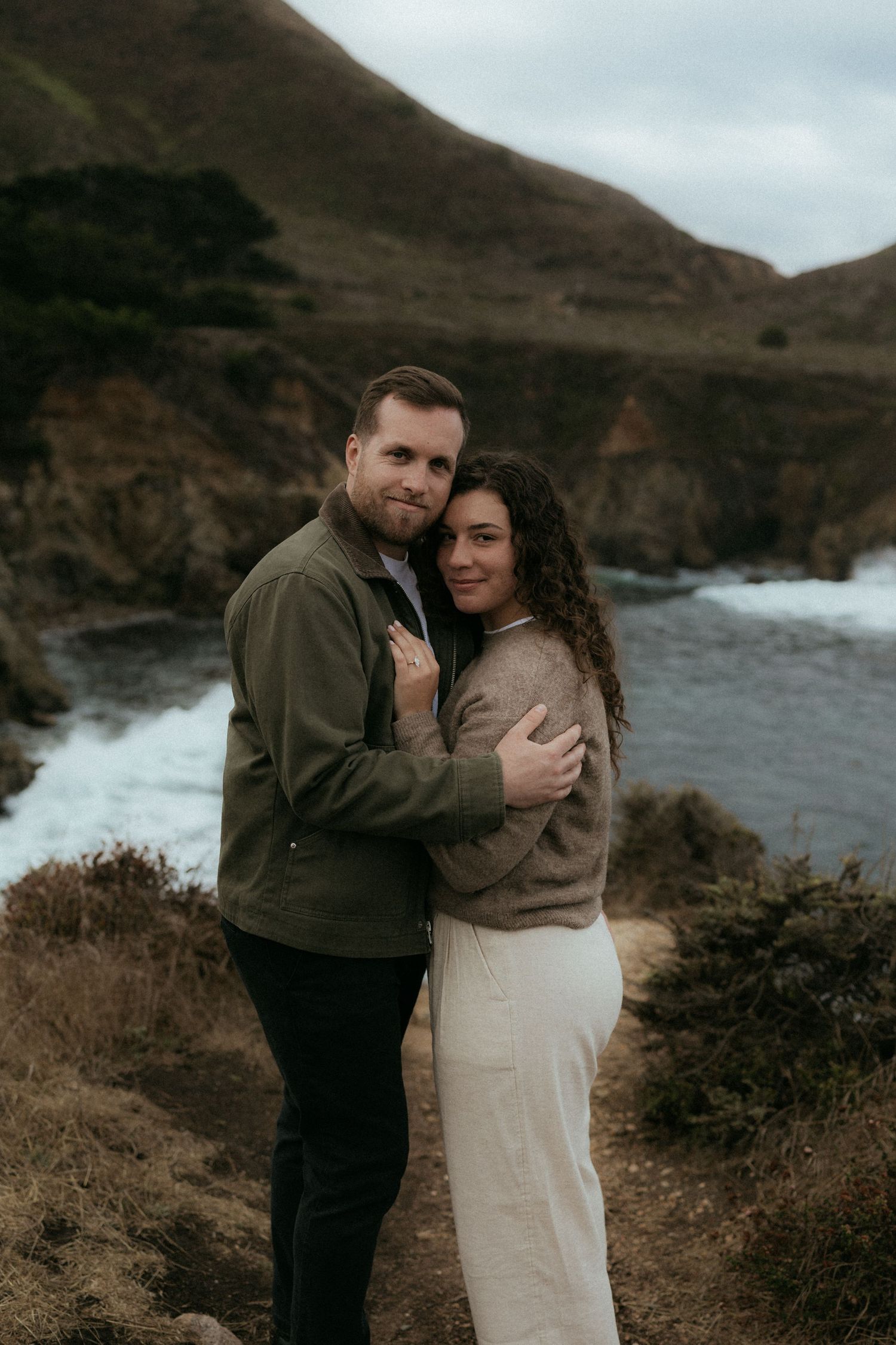 A couple embraces on a cliff overlooking a dramatic rocky coastline with crashing waves on an overcast day.