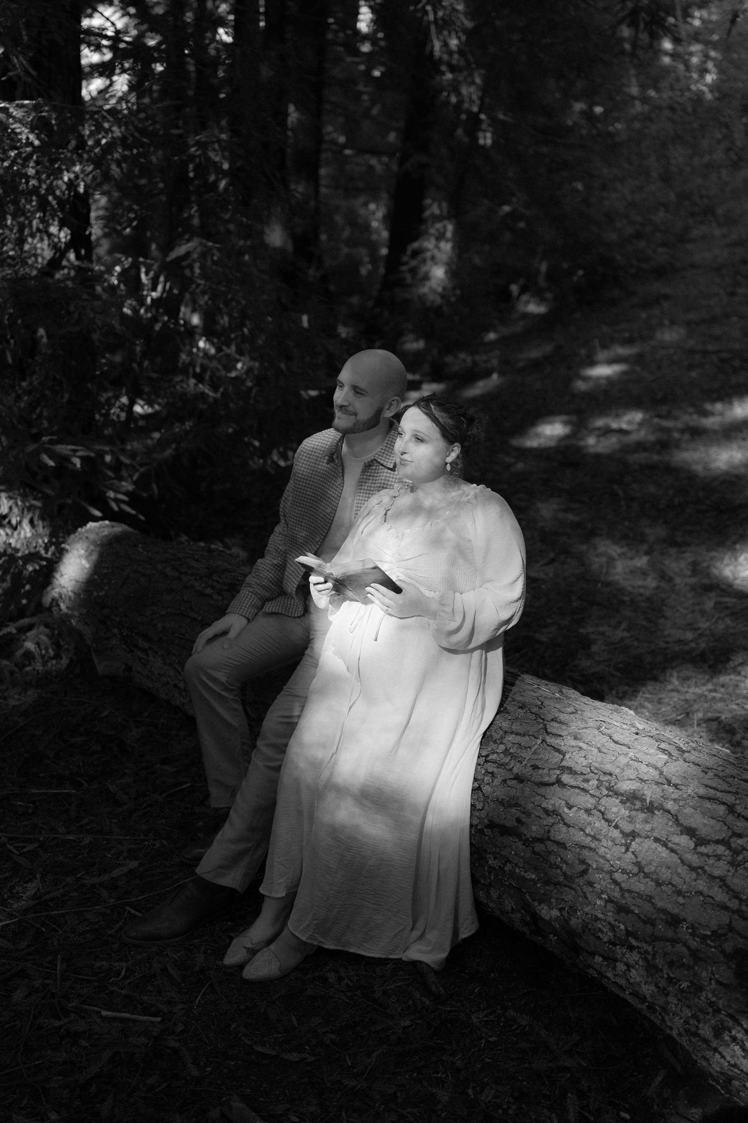 A couple sits together on a fallen log in a dreamy black and white forest setting.