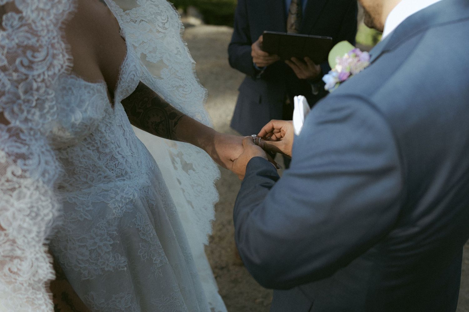Groom places wedding ring on bride's finger during outdoor ceremony, officiant holding book behind.