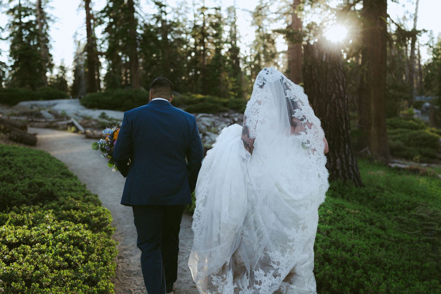 Bride and groom walking hand in hand through a sunlit forest garden path on their wedding day.