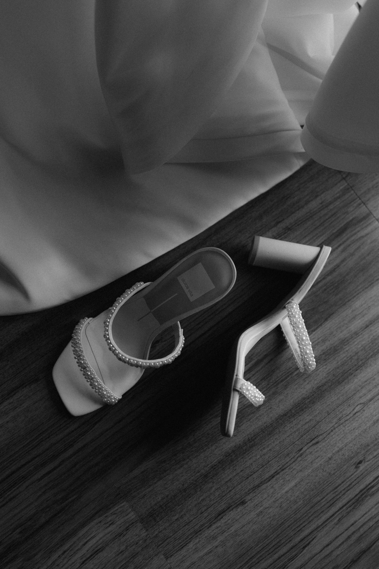 Black and white photo of elegant embellished heeled sandals on a wooden floor beside a flowing white dress.