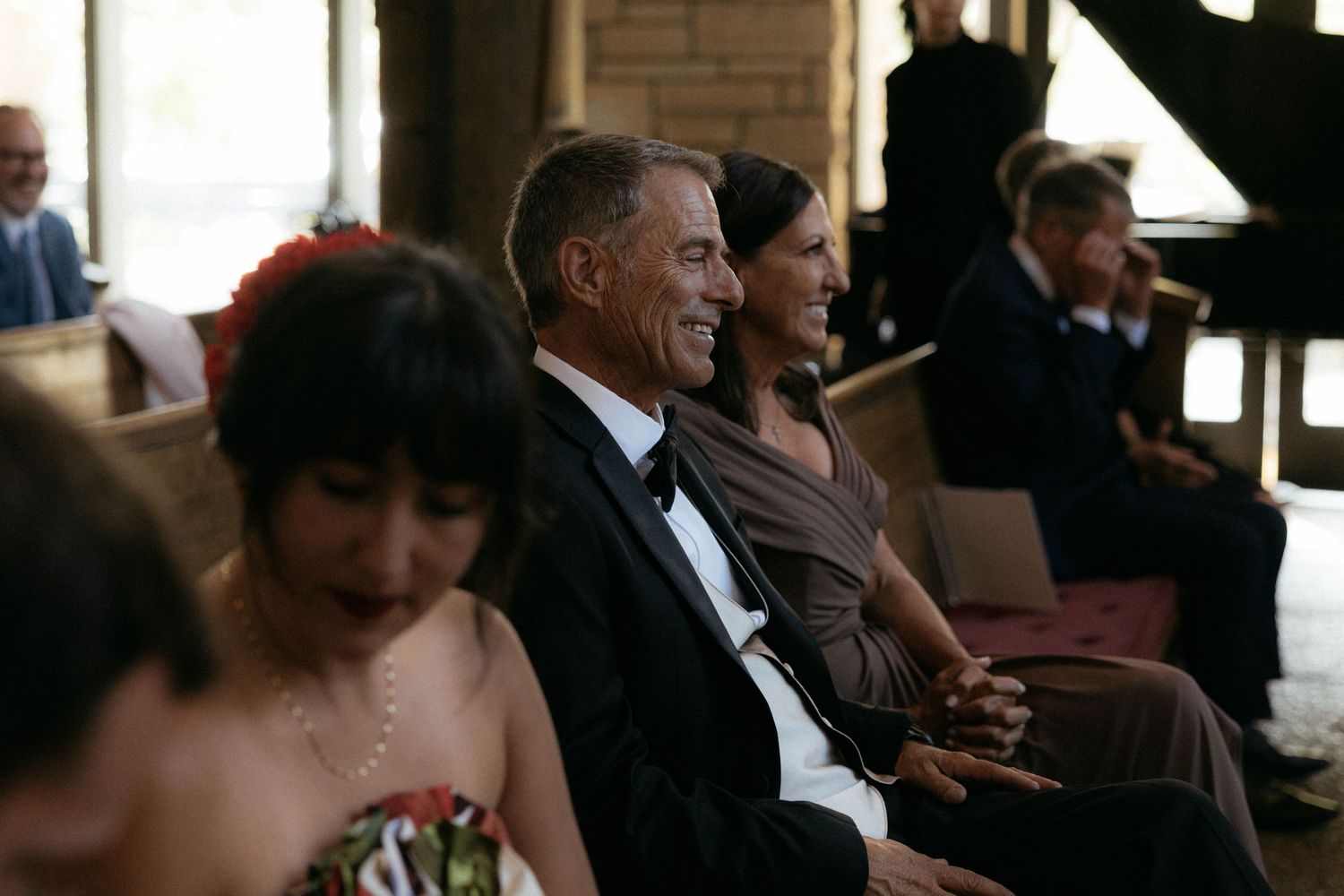 Smiling wedding guests seated in rows during an indoor ceremony, man in tuxedo and woman in brown dress.