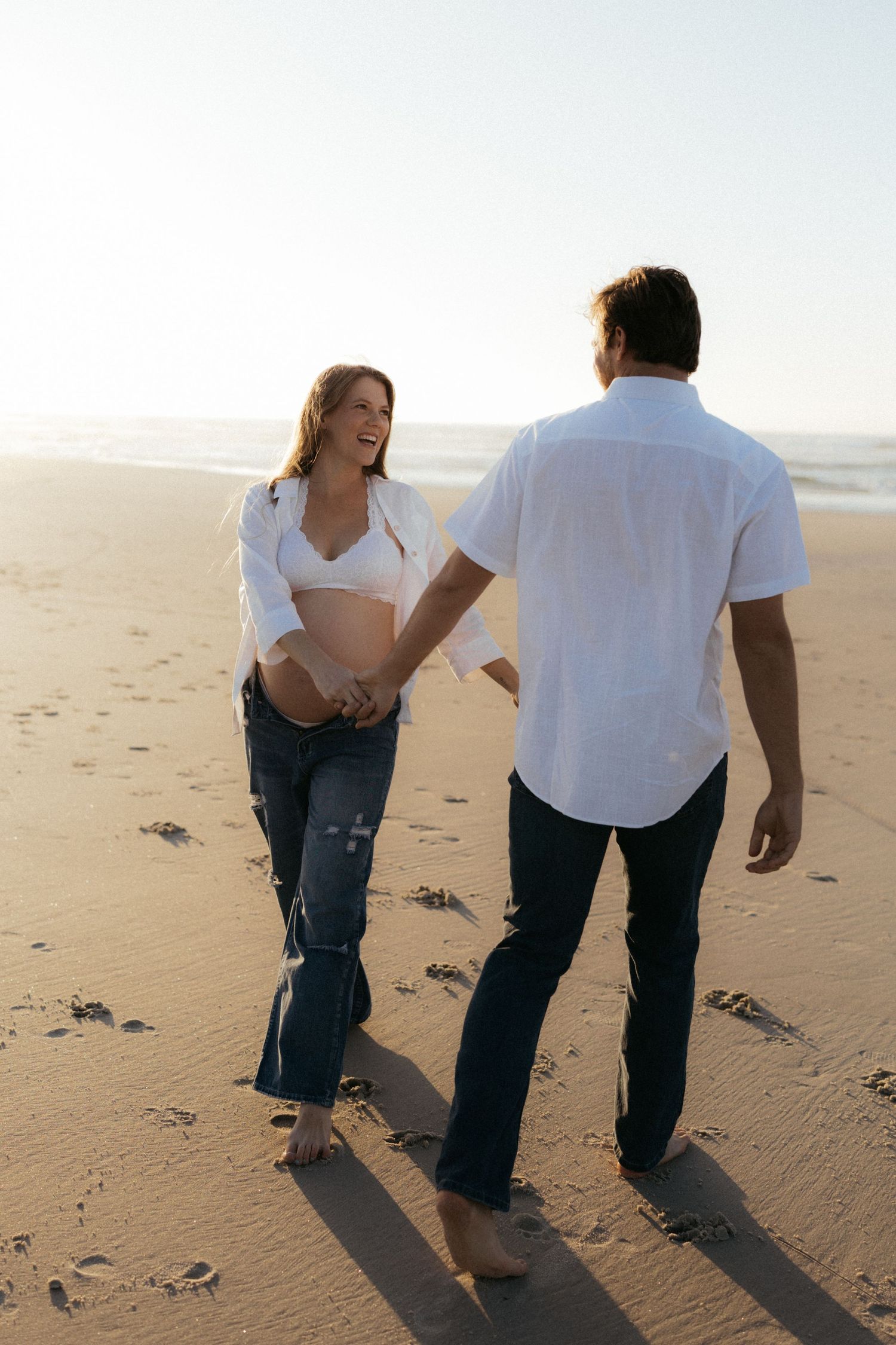 Pregnant woman holding hands with partner while walking barefoot on sandy beach at sunset.