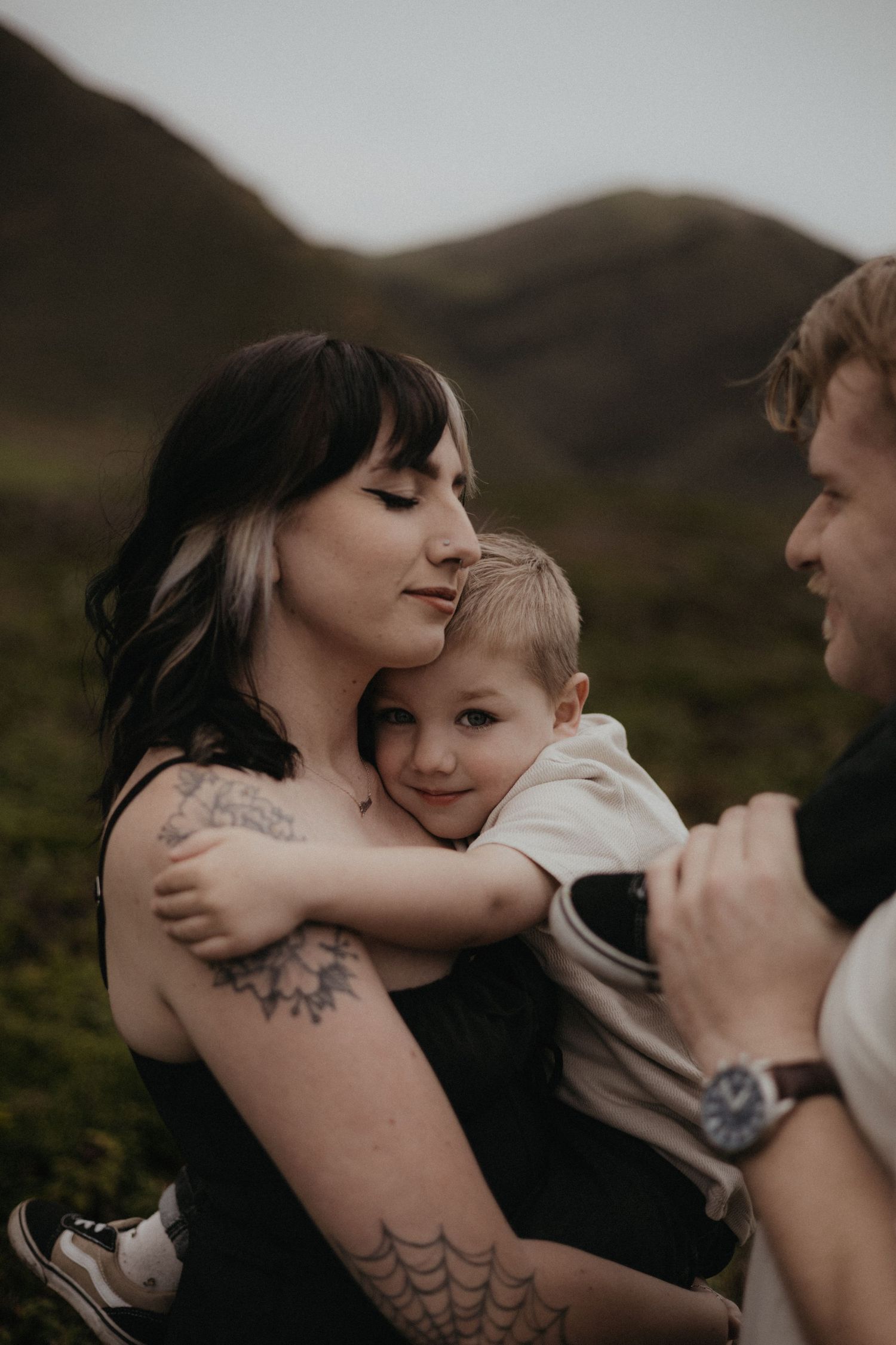 A tattooed mother holds her young son while a man looks on, set against a scenic mountain backdrop.