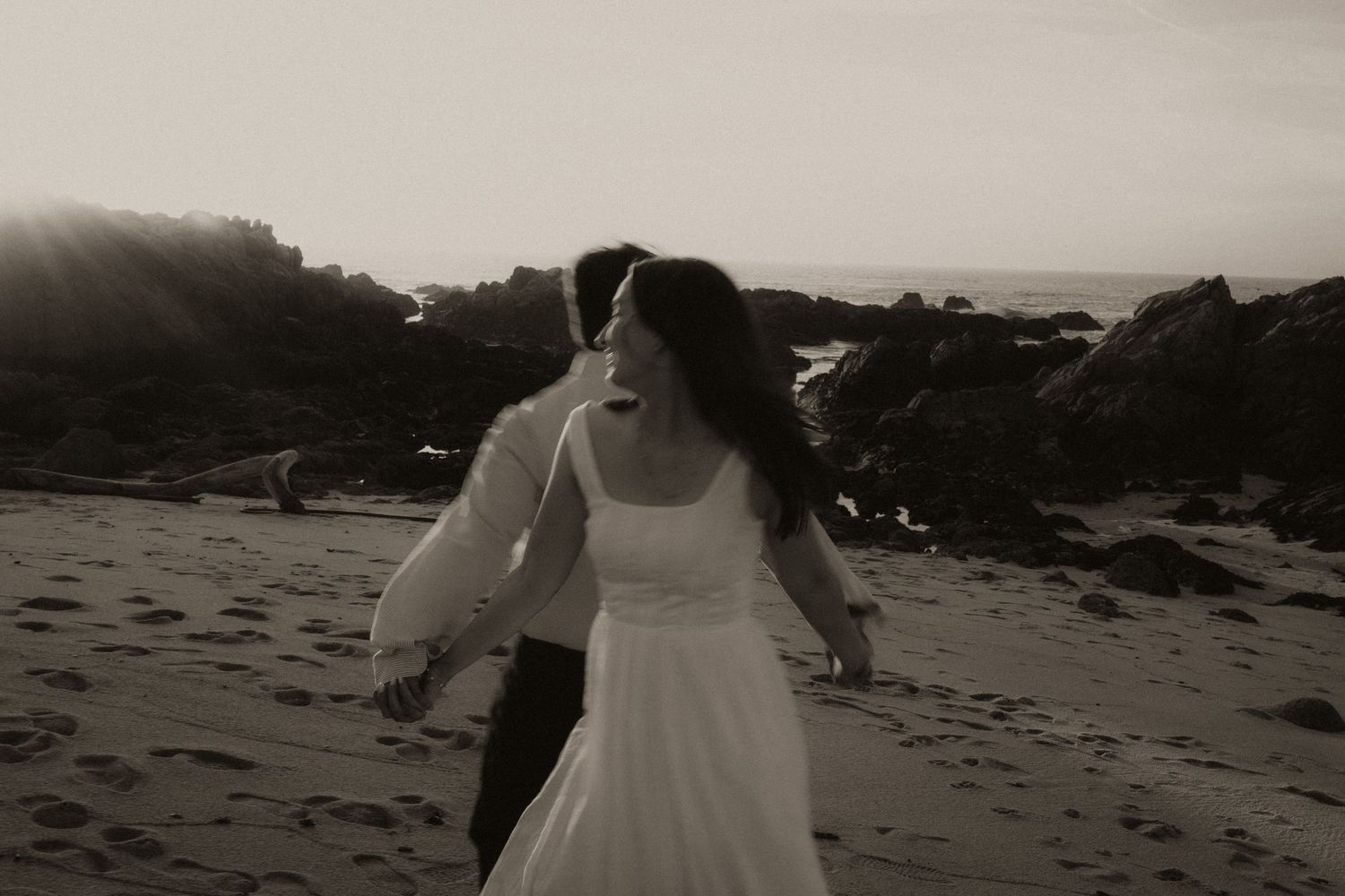 Black and white photo of a bride and groom walking on a rocky beach at sunset in wedding attire.