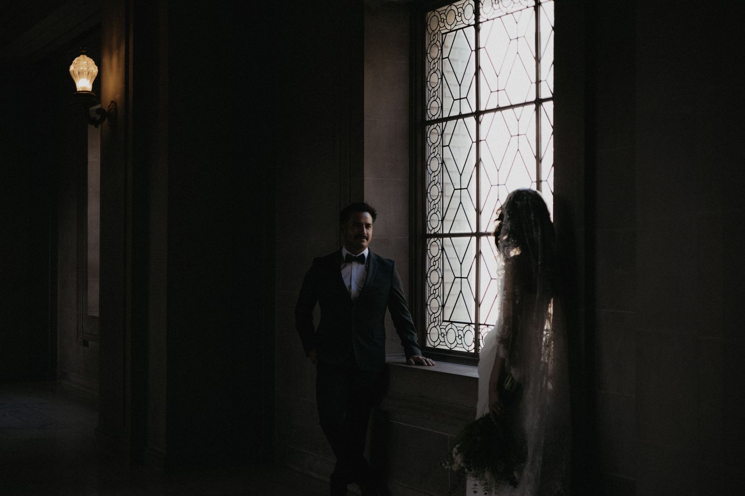 A moody silhouette of a bride and groom standing by a decorative window.