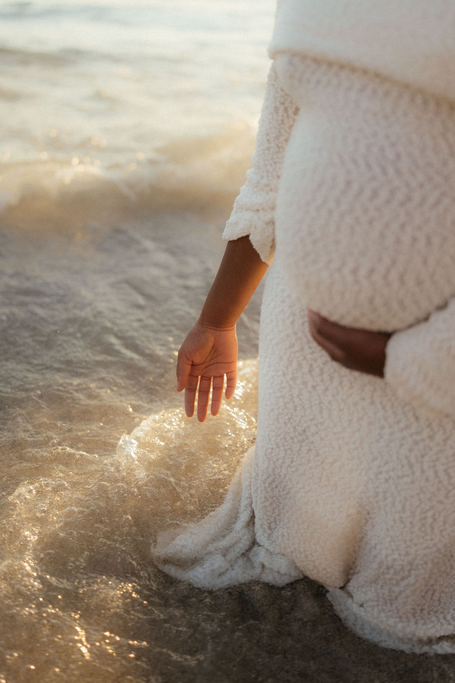 A person in a white lace dress dips their hand into glistening ocean waves during sunset.
