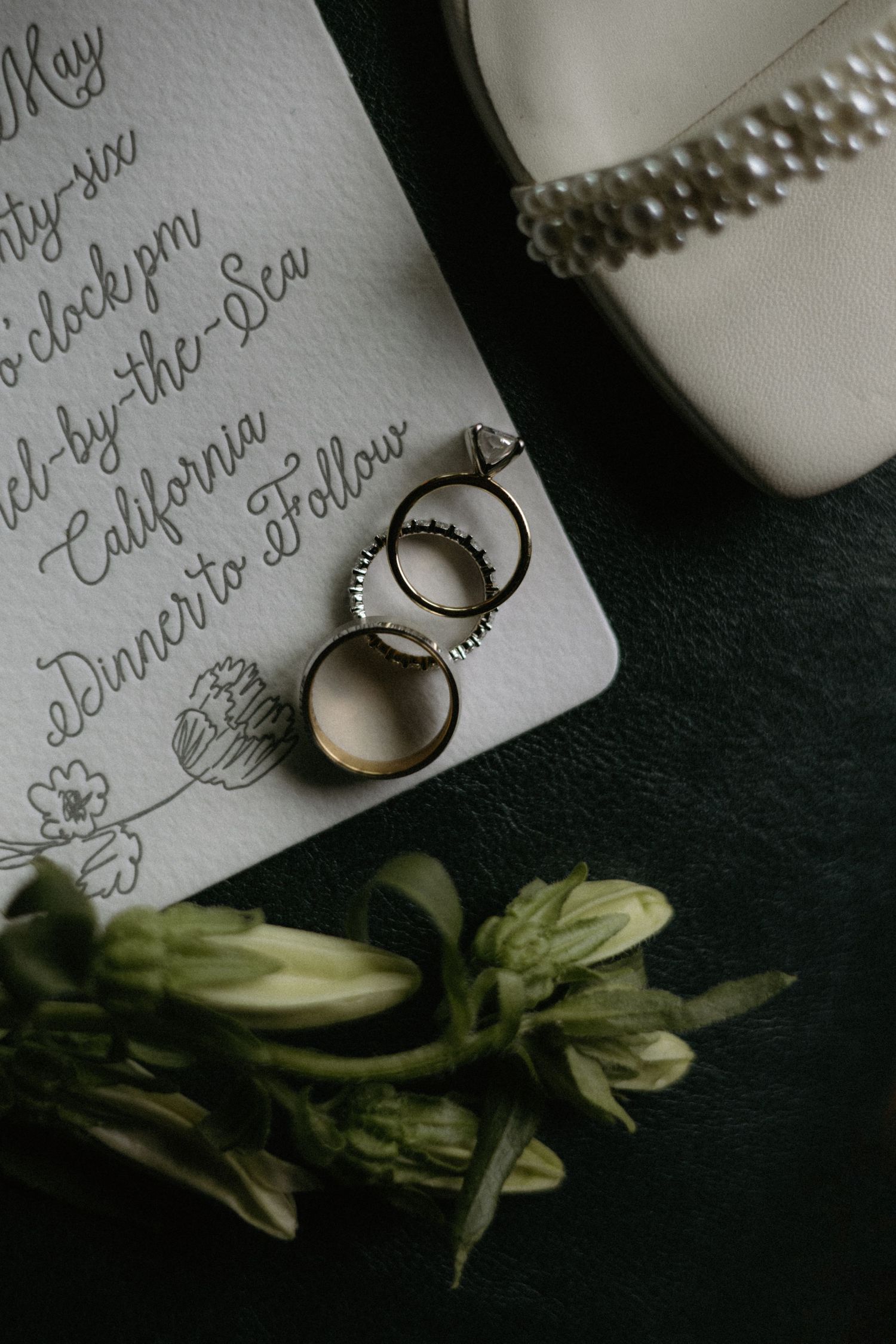 Wedding rings rest on a calligraphy invitation beside white floral buds and a pearl-adorned bridal heel on a dark surface.