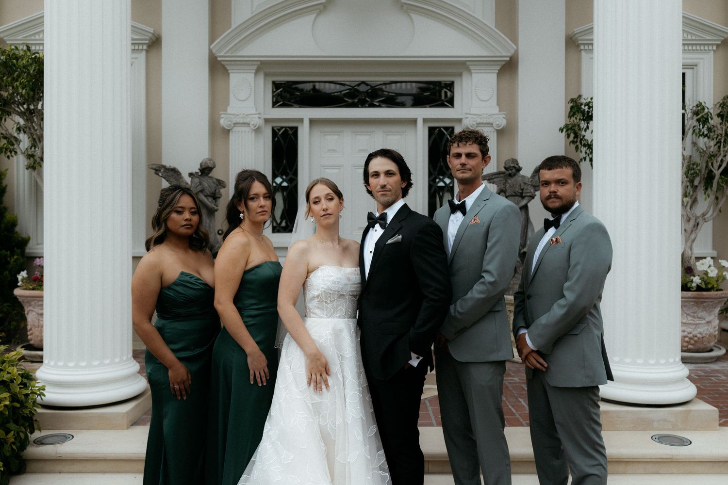 Wedding party poses before white columns, bride in white gown with groomsmen in tuxedos and bridesmaids in emerald green dresses.