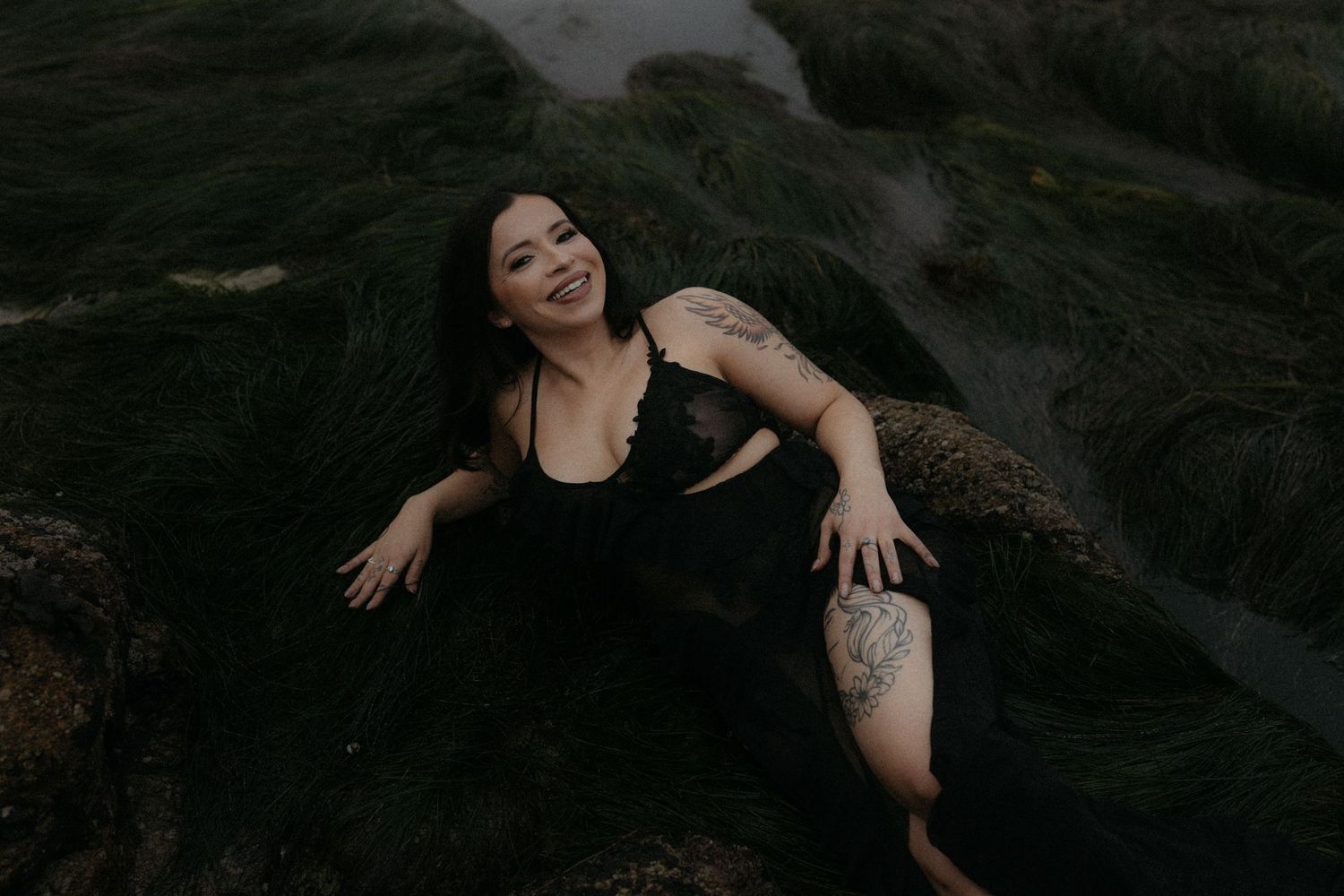 Woman in black dress smiling while reclining on moss-covered rocks outdoors, showcasing a floral tattoo on her thigh.