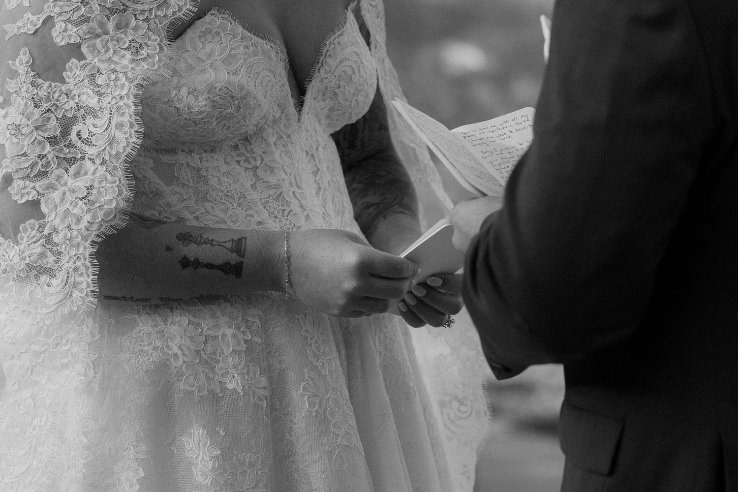 Black and white close-up of tattooed bride holding groom's hand during wedding ceremony.