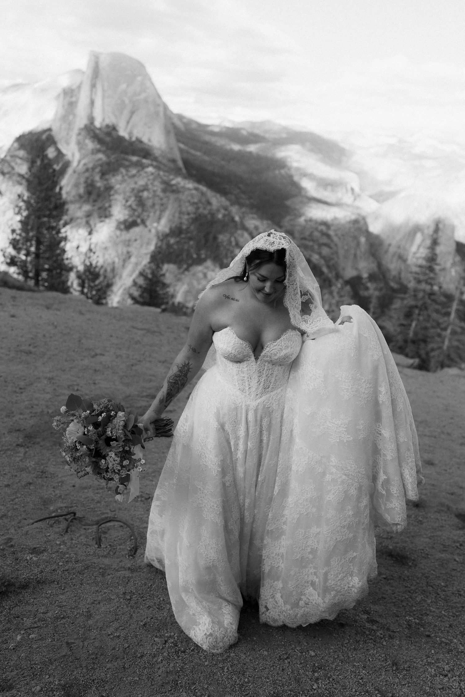 Bride in lace wedding gown and veil poses with wildflower bouquet at Yosemite with Half Dome in black and white photo.