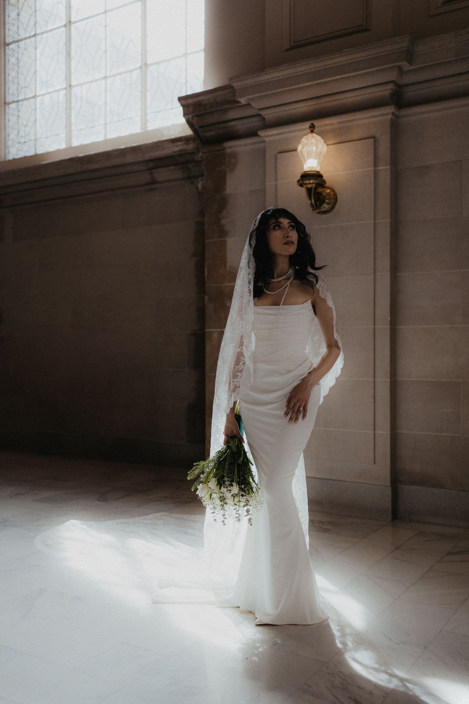 Bride in flowing white gown poses in dramatic window light creating ethereal shadows.