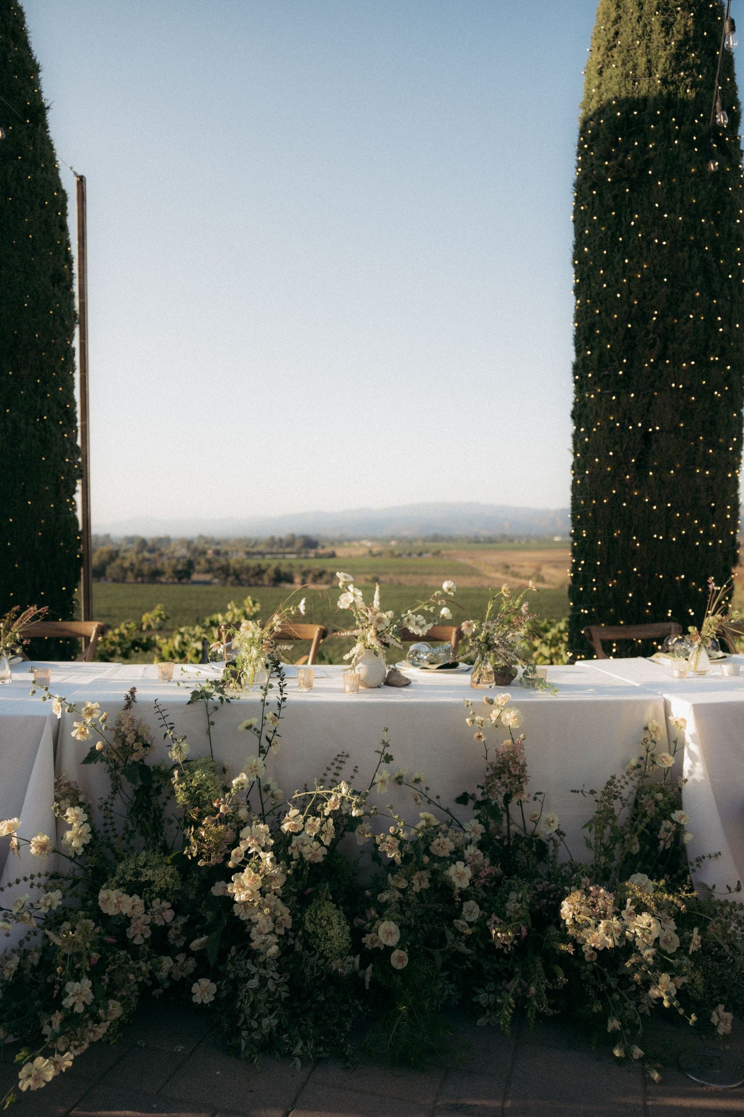 White garden roses and floral arrangements line a balcony wall with scenic countryside views.