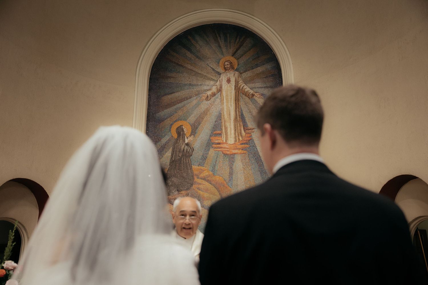 A religious wedding ceremony takes place in a church with a painted mural of Jesus visible in the background.