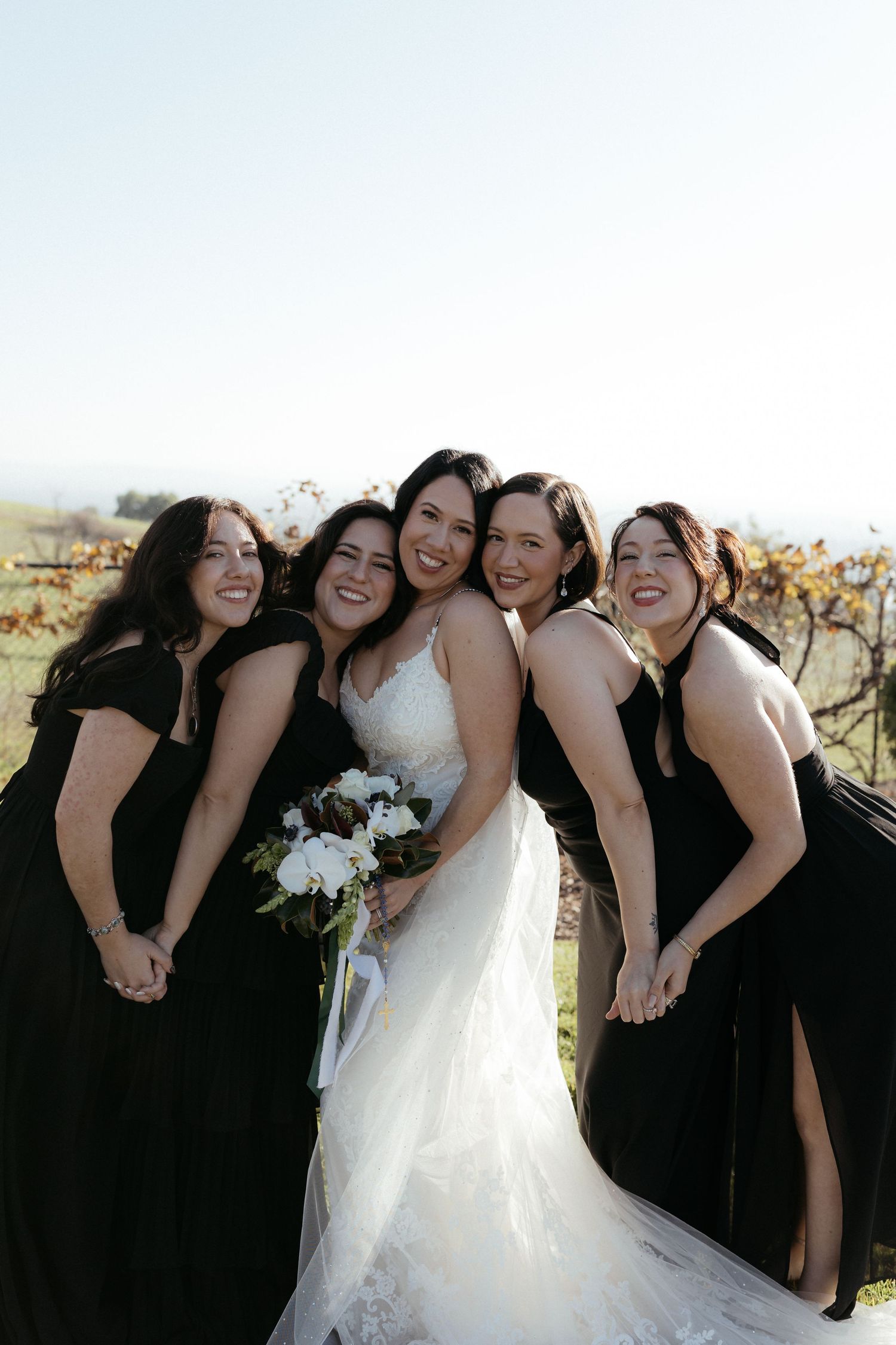 A joyful wedding party poses together with bridesmaids in black dresses surrounding the bride in white holding a bouquet.