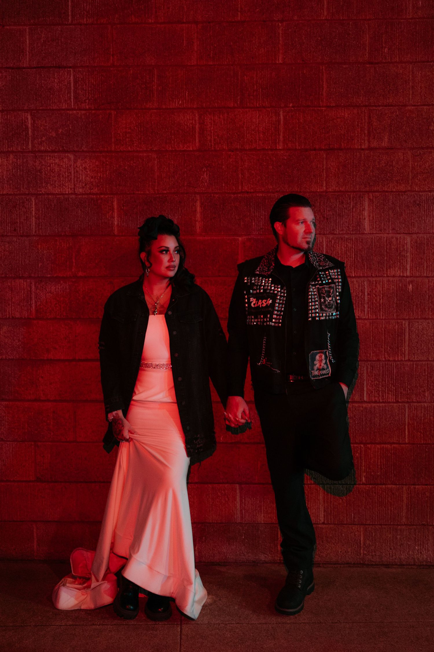 A stylish couple poses against a red-lit brick wall, combining formal and punk fashion elements.