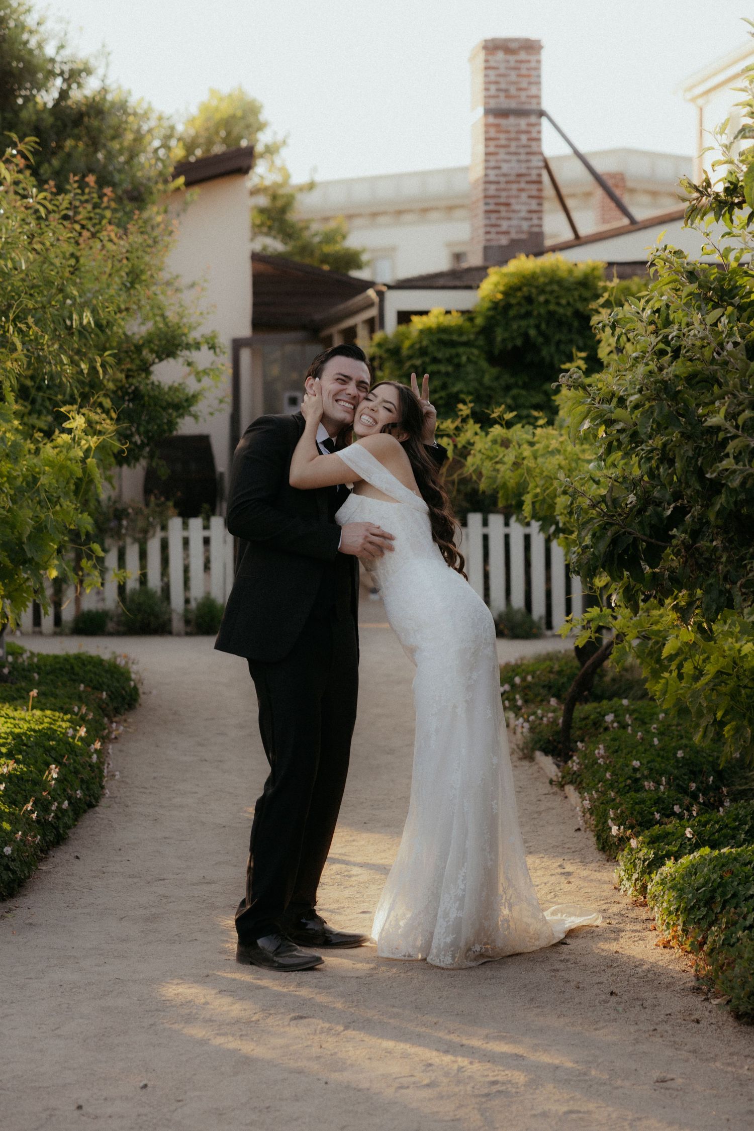 A couple embraces on a garden path with a white picket fence and cottage in the background.