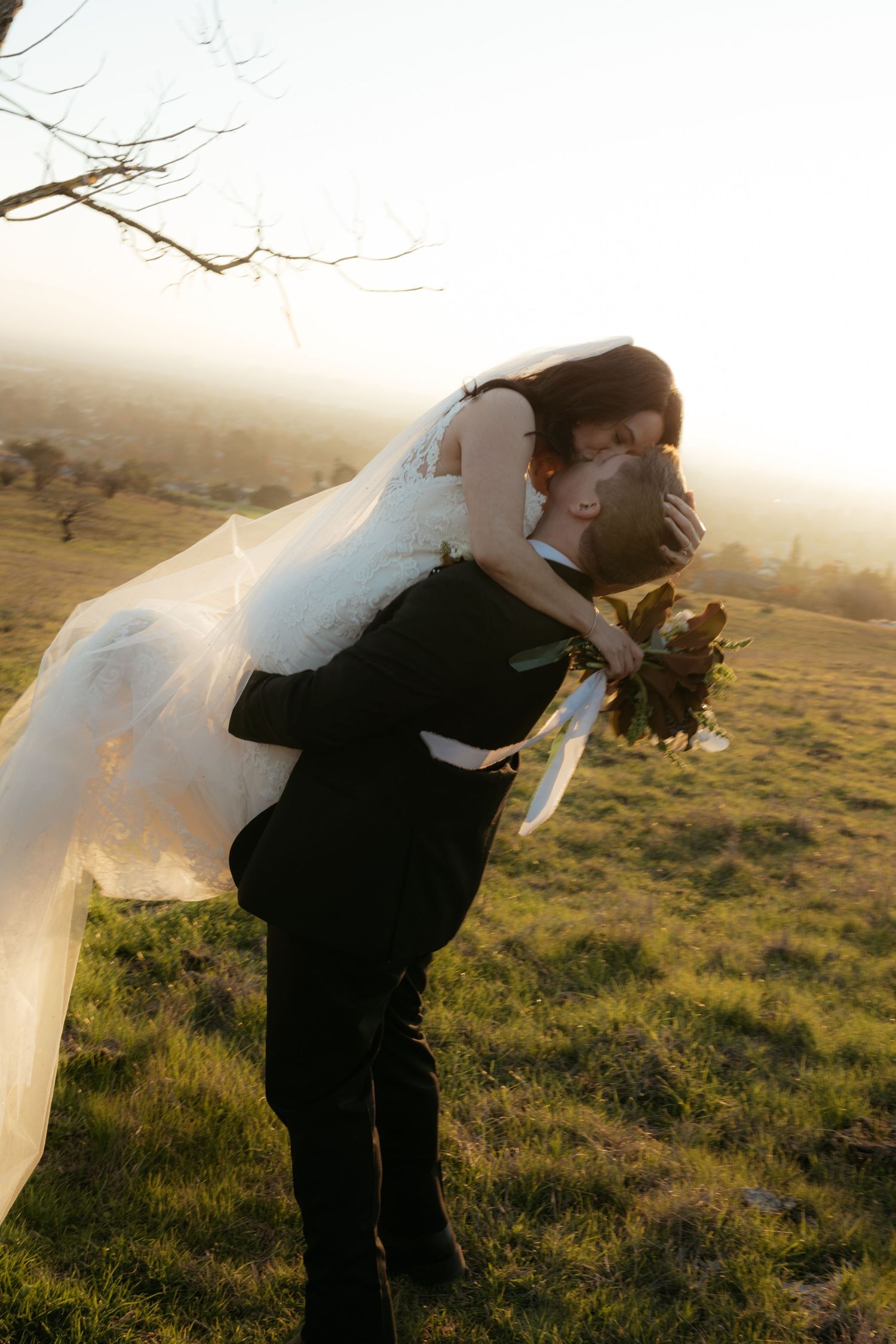 A romantic wedding couple embraces in a dramatic dip pose against a golden sunset landscape.