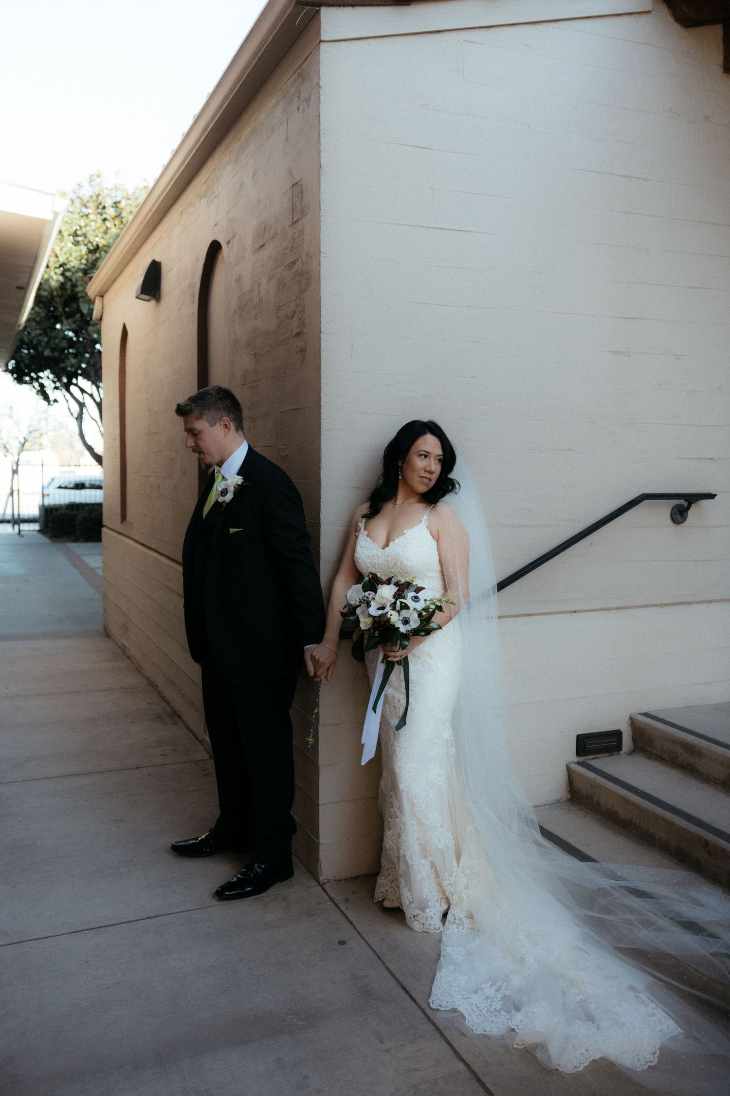A couple poses separately against a beige wall before their wedding ceremony, with the bride holding a bouquet.